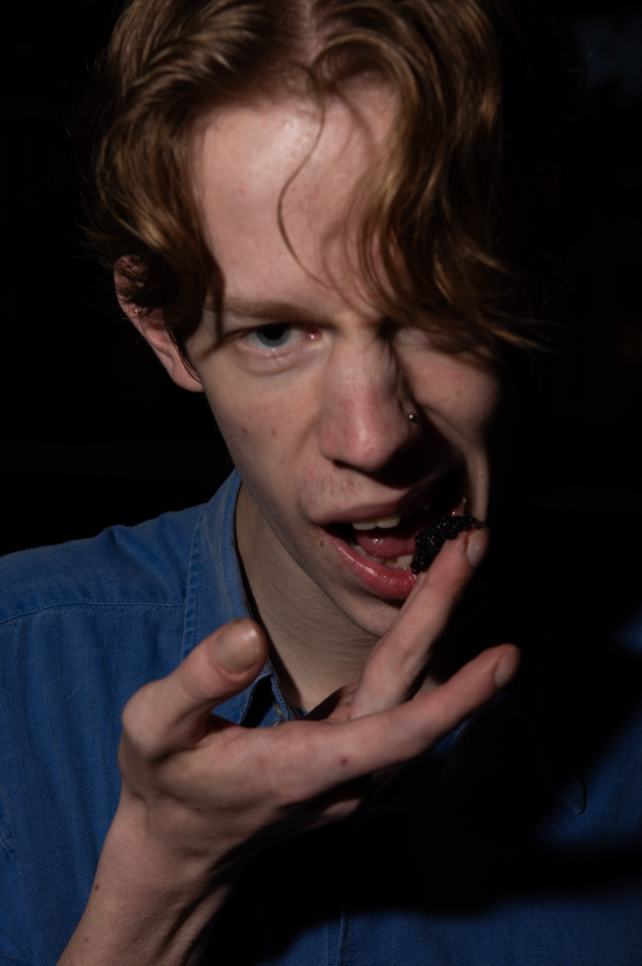 A person with red hair and a nose piercing eating a blackberry, wearing a blue shirt against a black background.