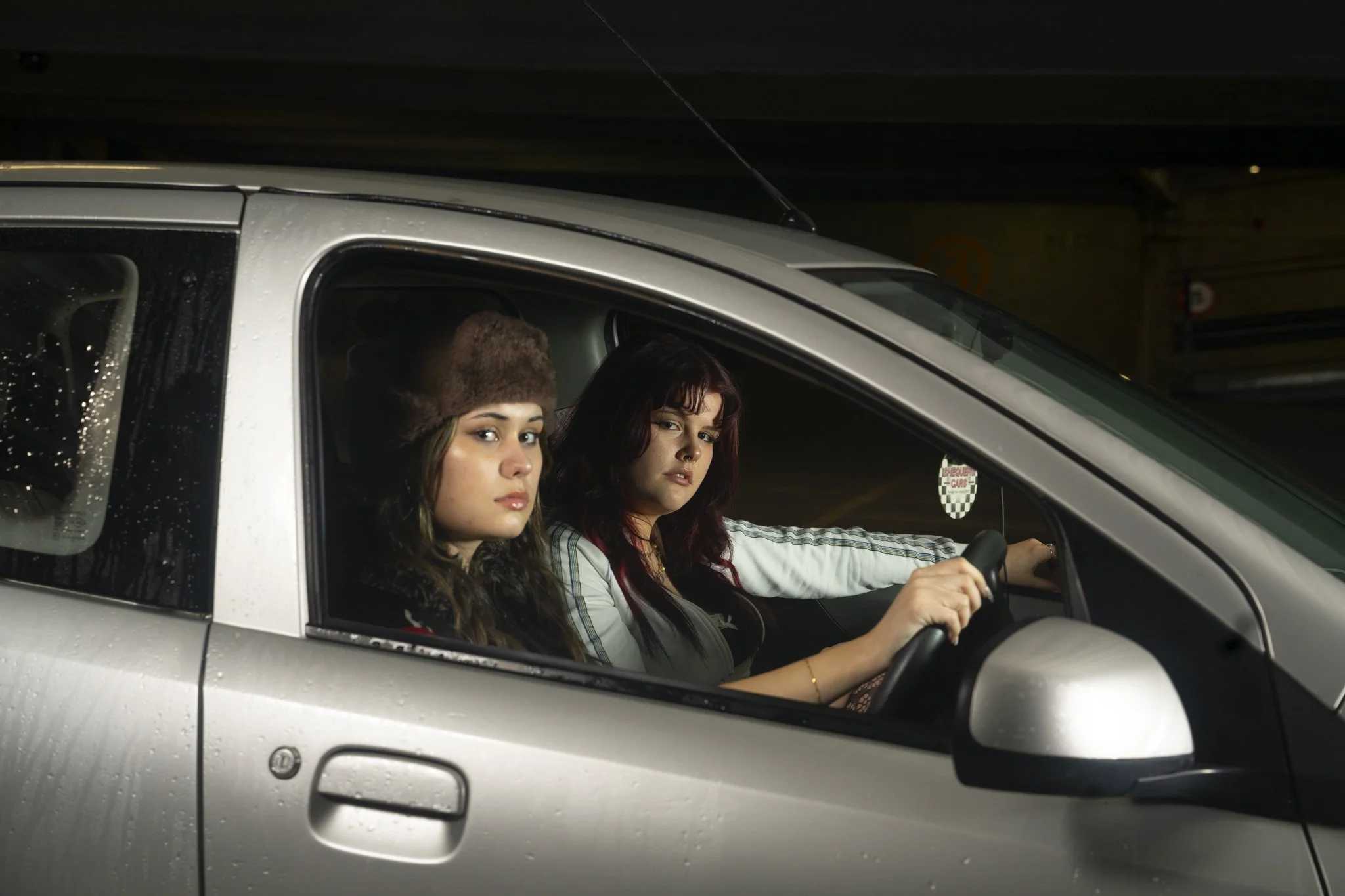 Two women sitting in a silver pickup truck at night, with one woman driving and the other looking out the window.