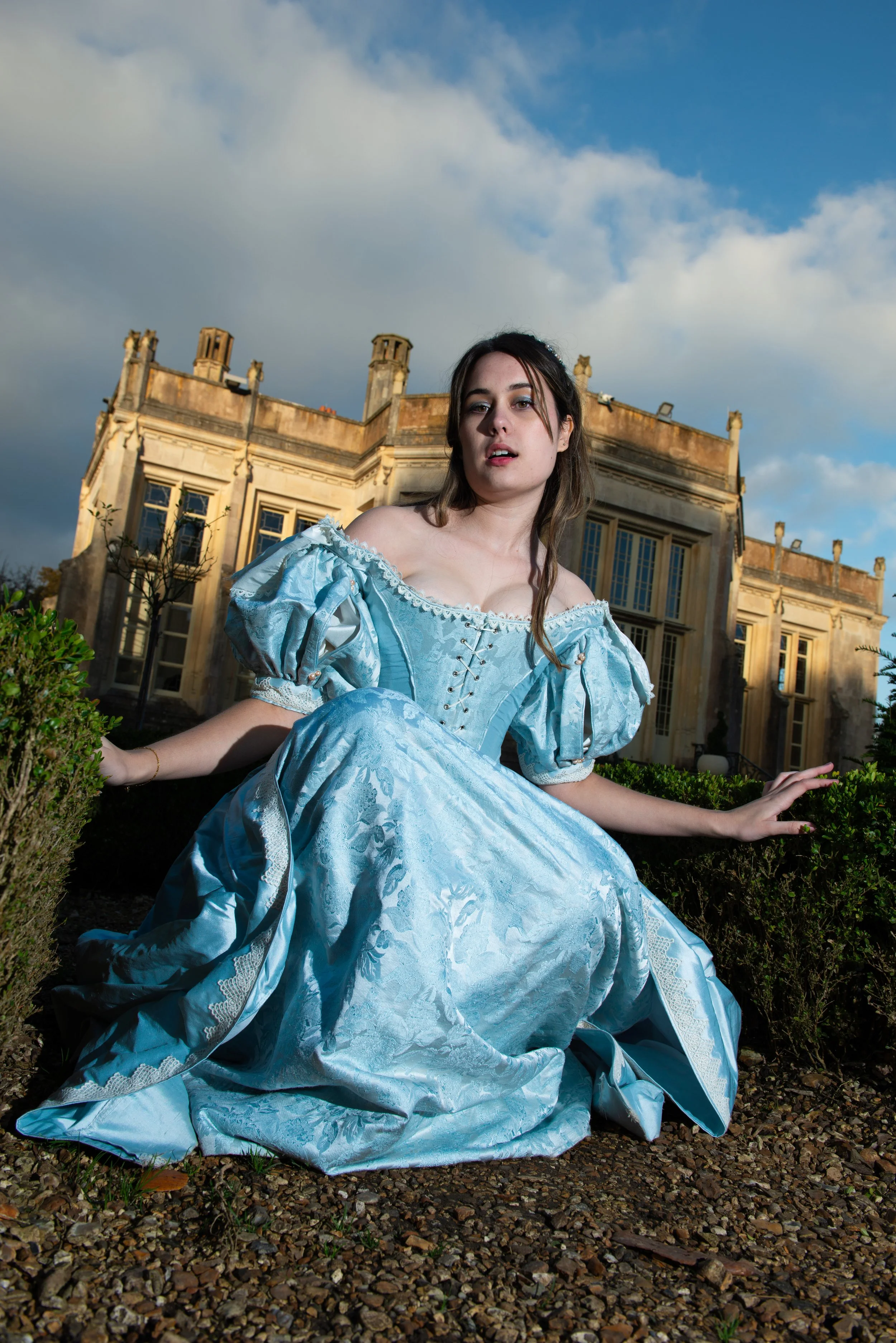 A woman in a blue historical dress sitting outdoors on gravel in front of a large, old building with a tower and cloudy sky.