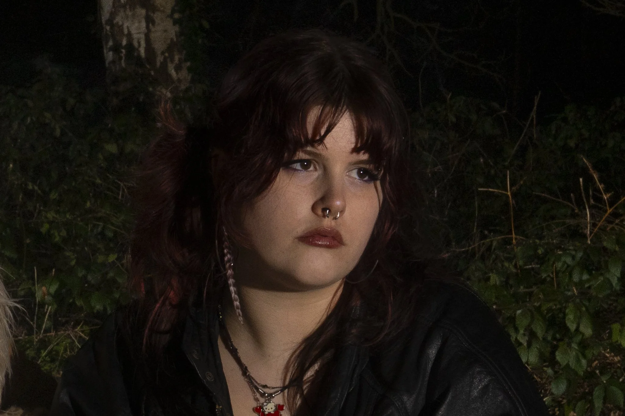 A young woman with dark red, wavy hair and makeup, featuring bold eyeshadow and lipstick. She has facial piercings, including a septum ring, and is wearing necklaces, one with a Hello Kitty pendant. She is outdoors at night, with dark, leafy foliage in the background.
