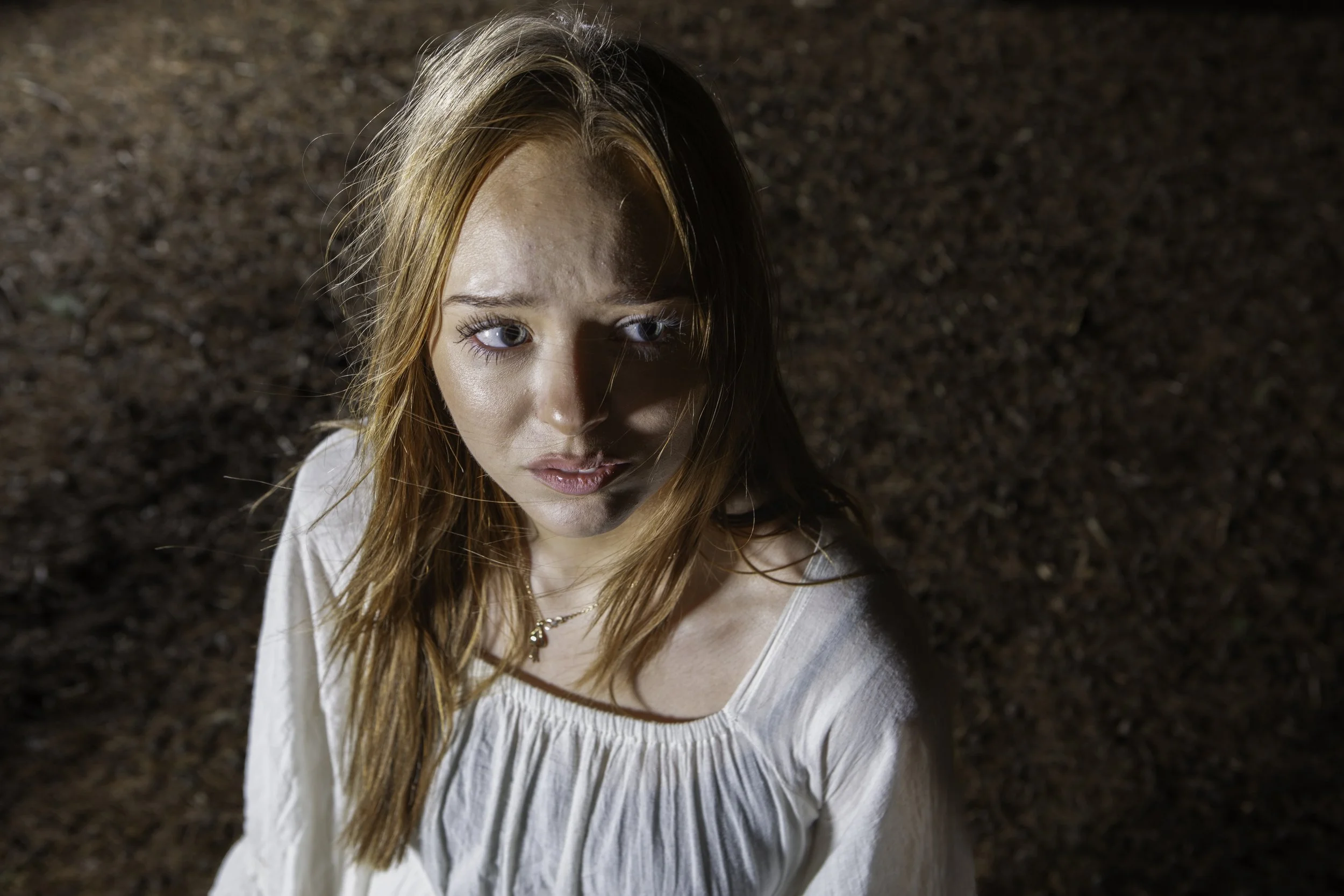 A young woman with long red hair and blue eyes stands outdoors on a dirt surface. She is wearing a white, loose-fitting top and a necklace, looking up towards the camera with a neutral expression.