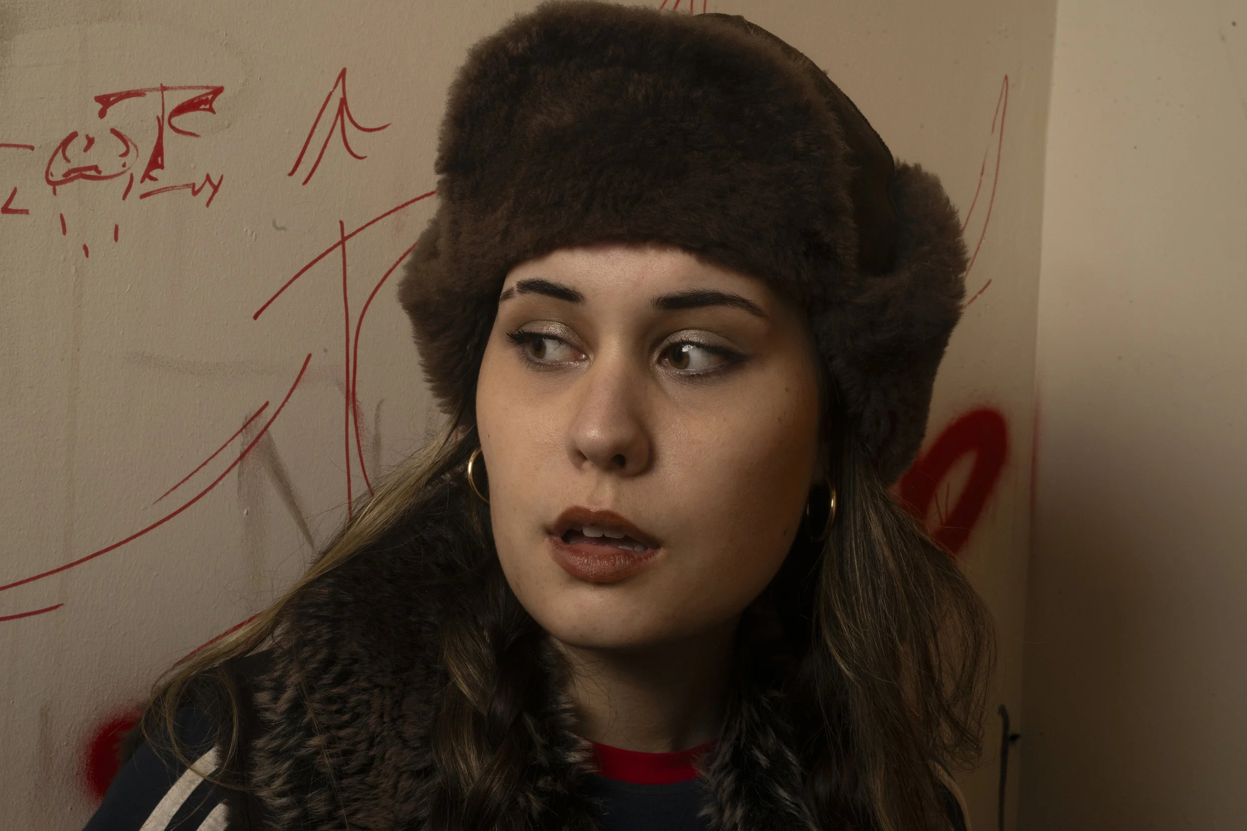 A woman with long, wavy hair wearing a fluffy, dark brown hat and a fur jacket, standing in front of a wall with red graffiti.