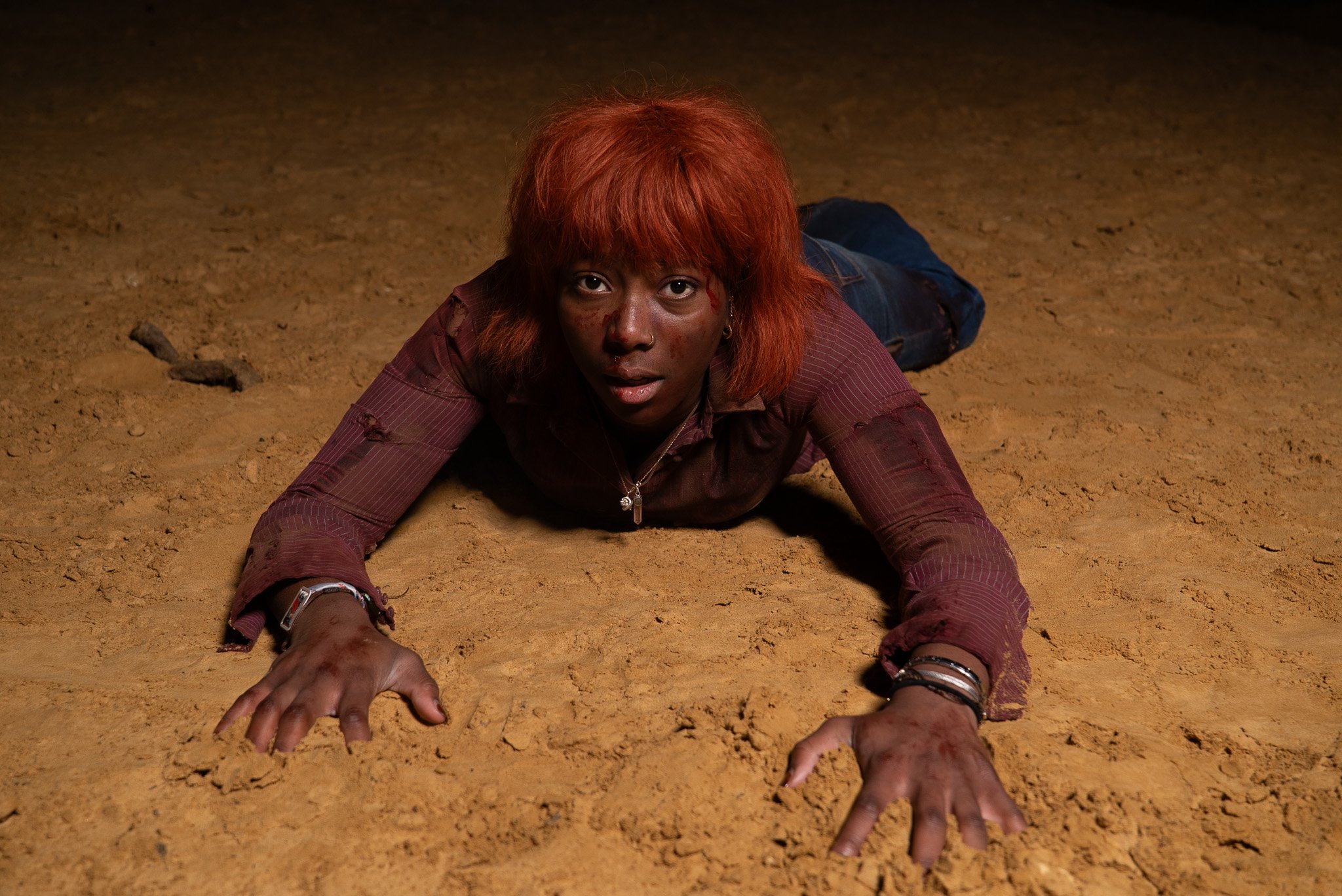 A woman with bright red hair lying on the ground, looking directly at the camera with her hands stretched forward on the dirt surface.