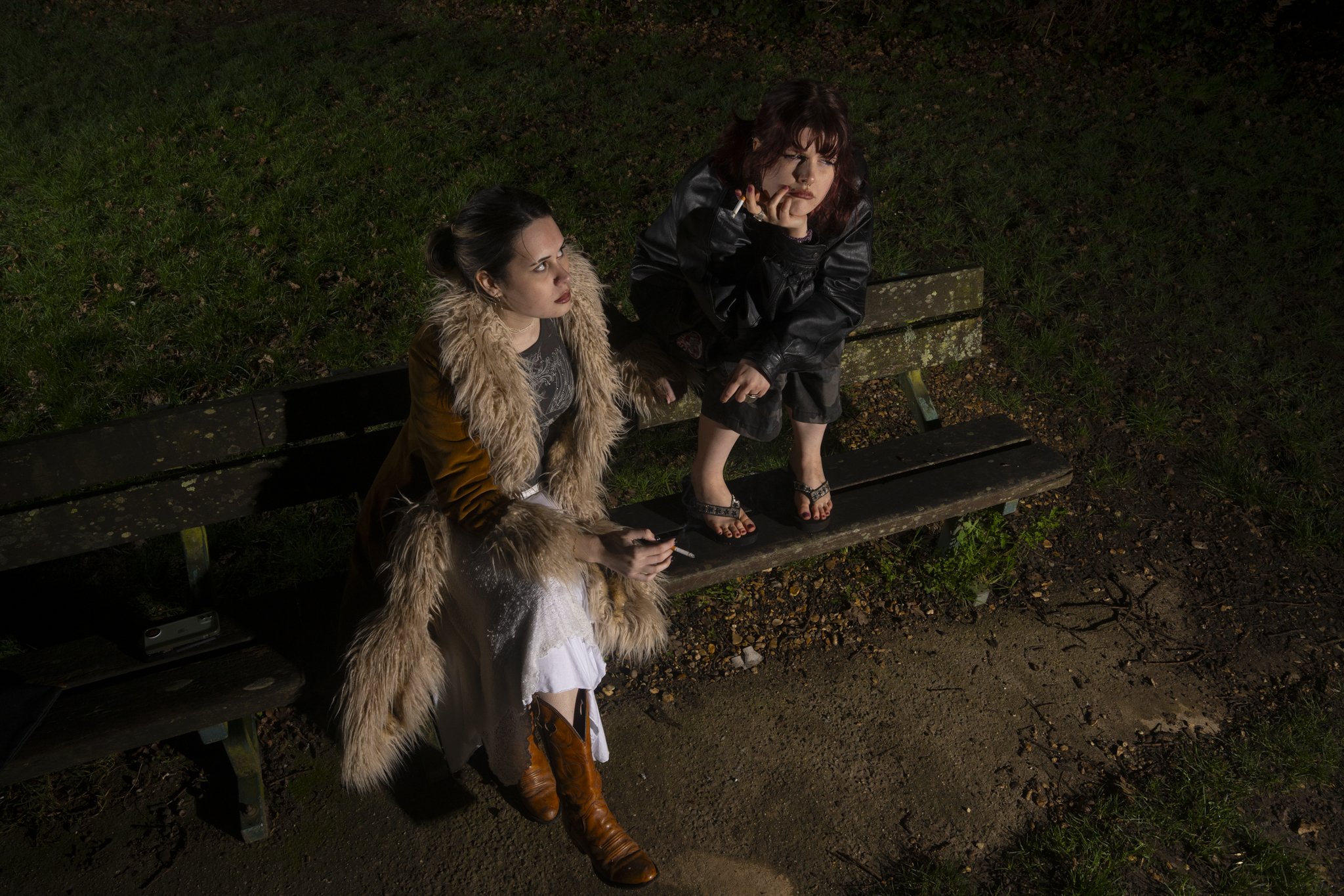 Two young women sitting on a park bench at night. One woman has dark hair in a bun and is wearing a fur-lined brown jacket, the other has wavy brown hair and is wearing a black leather jacket. Both are smoking cigarettes and looking in different directions.