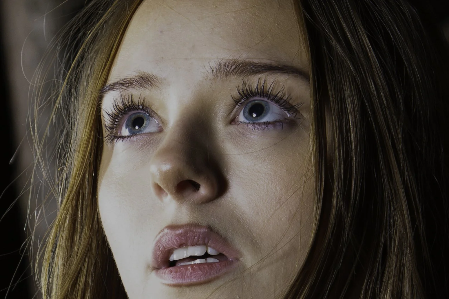 Close-up of a young woman with blue eyes, red hair, and makeup, looking slightly upward with a surprised or curious expression.