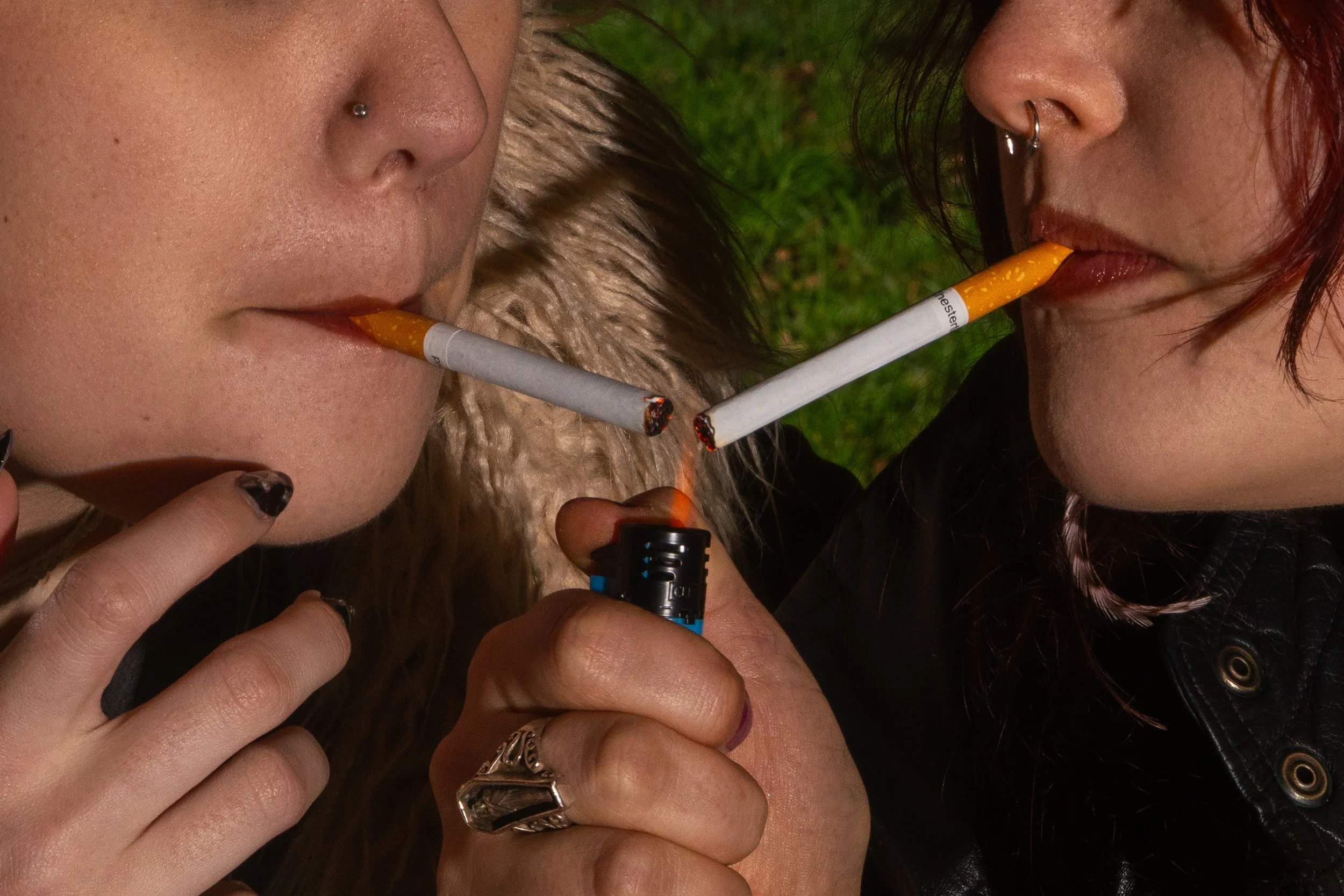 Two women smoking cigarettes outdoors on grass, one with a nose ring and the other with a septum piercing, both with dark hair.