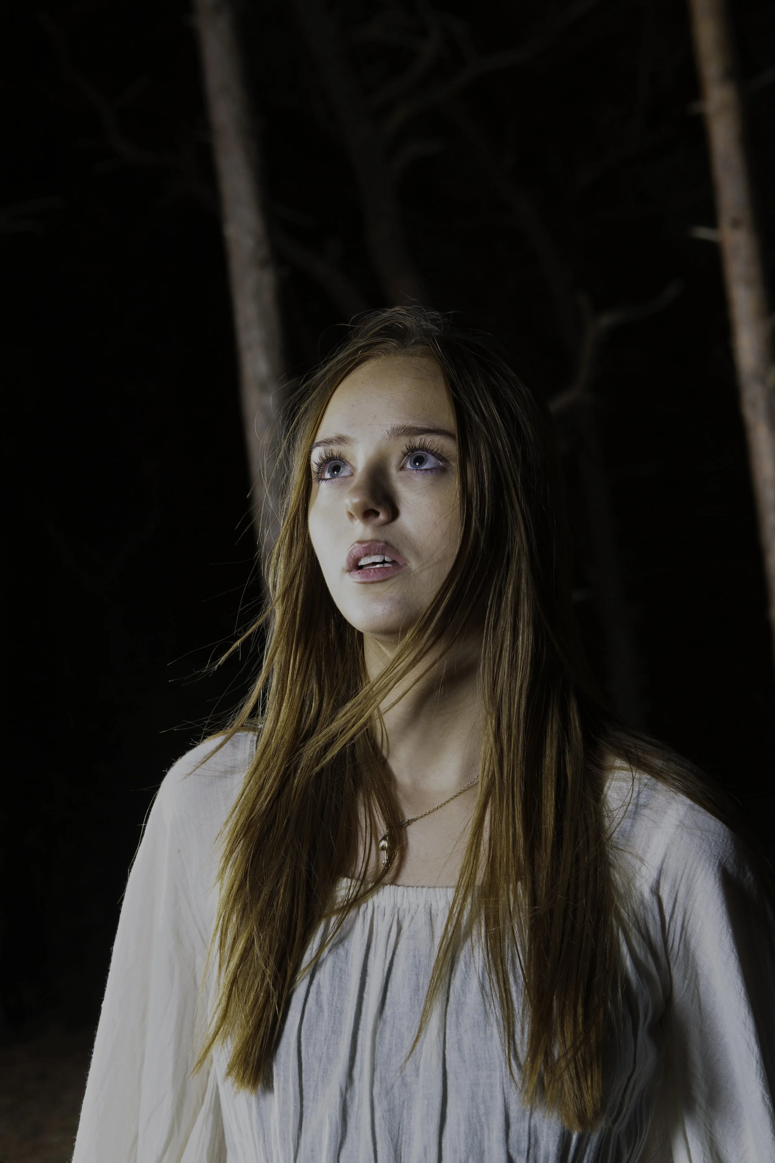 A young woman with long, red hair and blue eyes looking upwards, wearing a white blouse with thin vertical stripes and a gold necklace, standing outdoors at night with dark trees in the background.