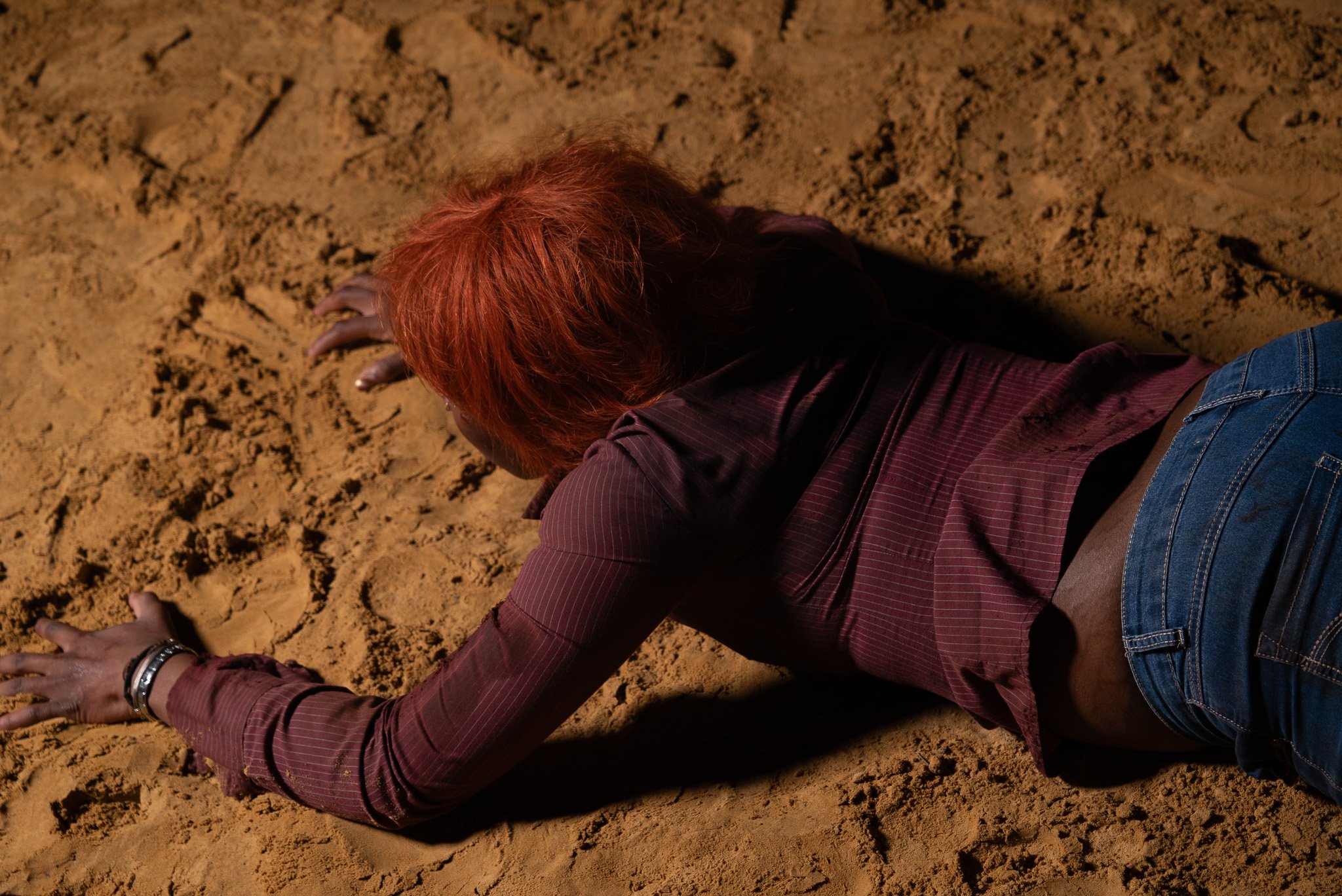 A person with red hair wearing a maroon shirt and blue jeans, lying face down on sandy ground with hands spread out.