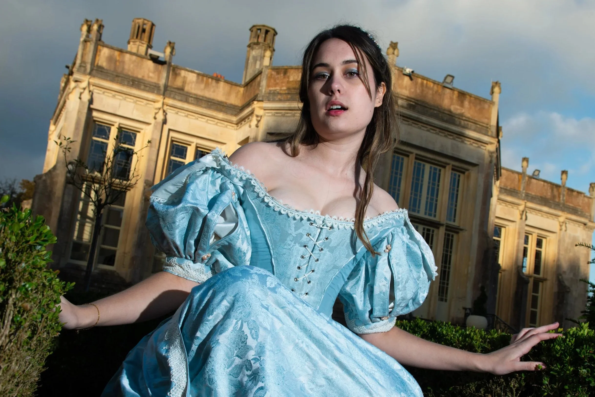 A woman in a light blue, Victorian-style dress with lace and puffed sleeves, posing outdoors in front of a historical building with large windows and a decorative roofline, under a partly cloudy sky.