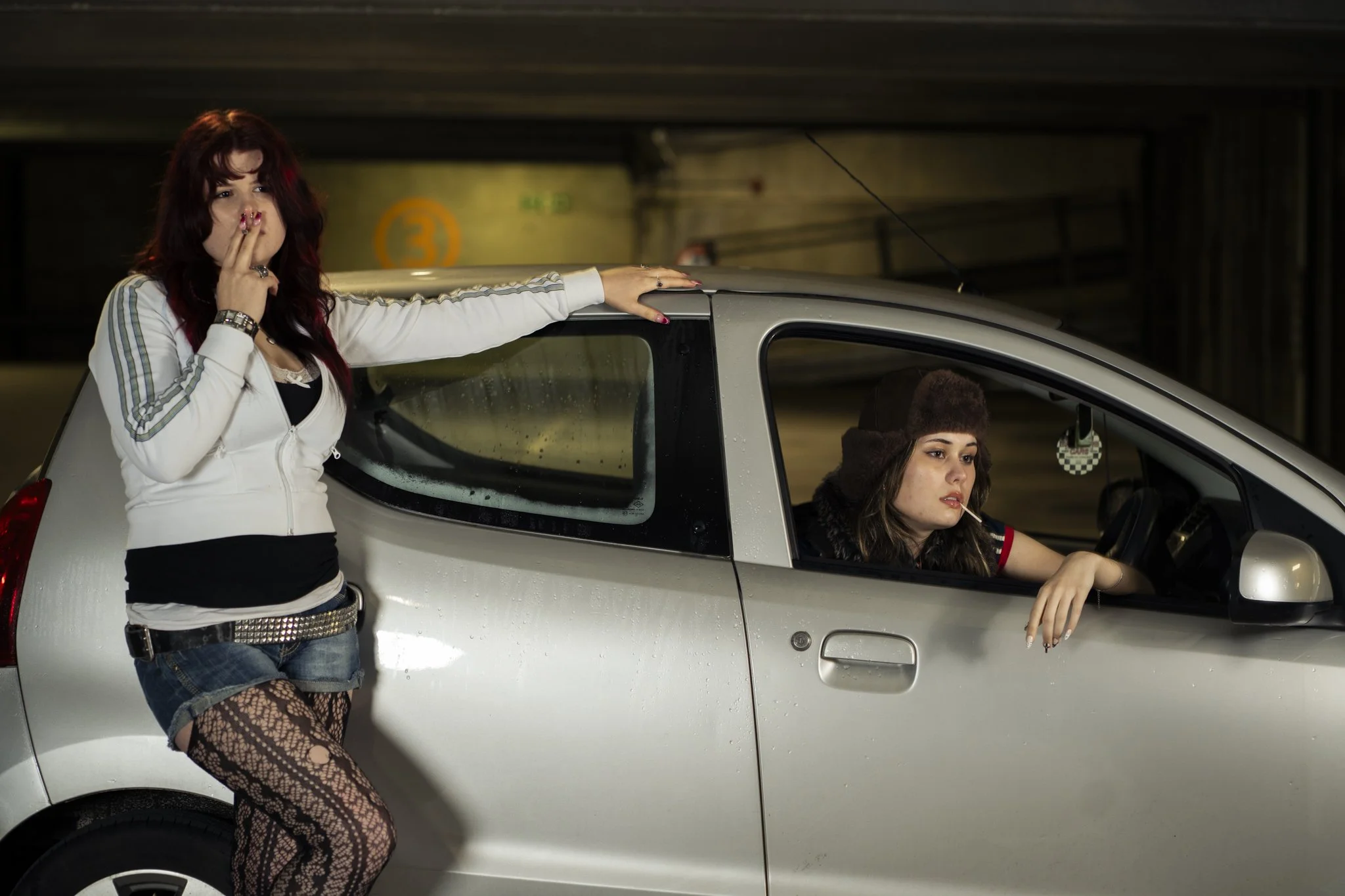 Two women with dark hair, one standing with her hand on a silver car, and the other sitting inside the car, in a parking garage. The woman standing wears a white jacket, shorts, and fishnet stockings, while the woman in the car has a hat and a lollipop.