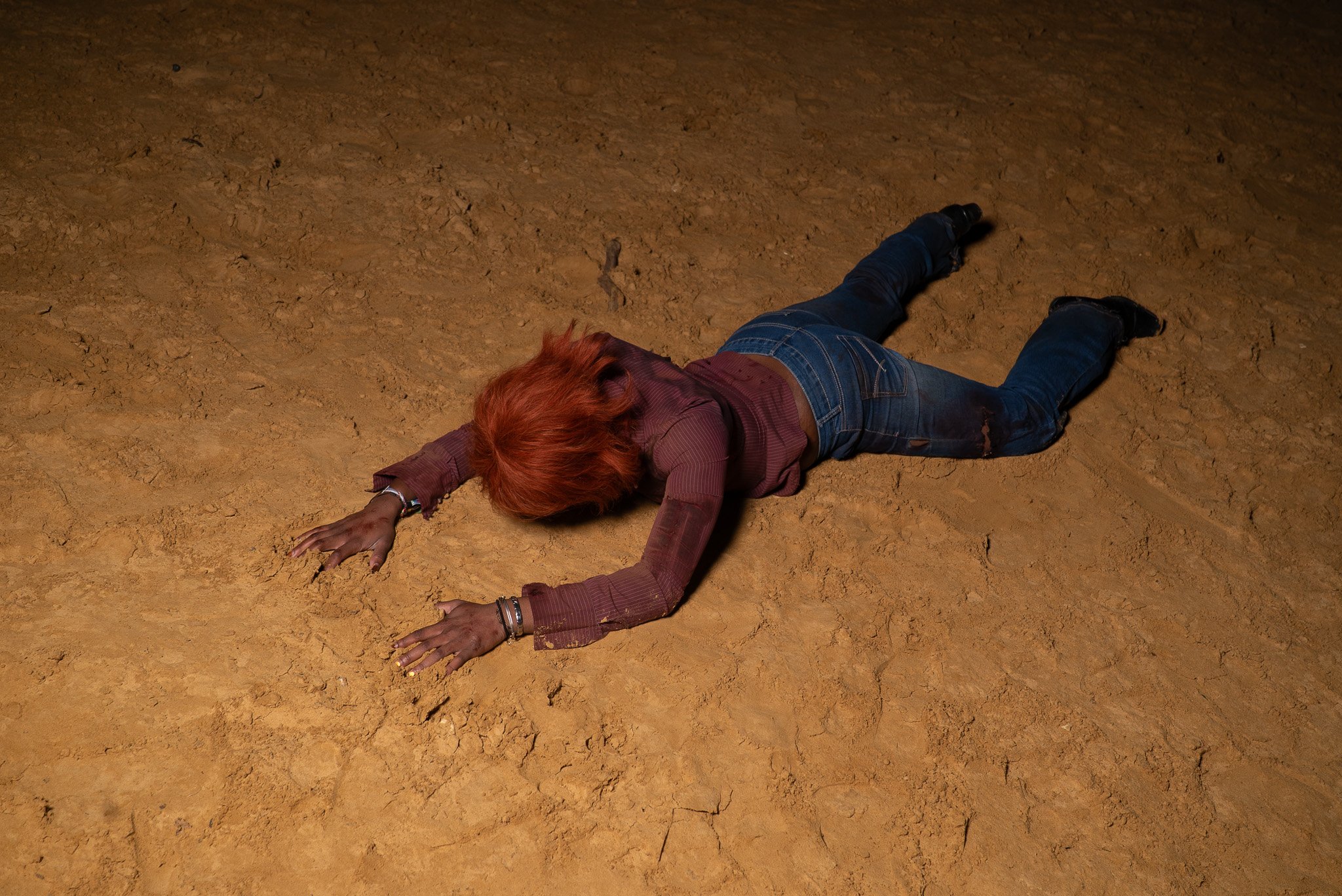 A woman with red hair lying face down on a cracked, dry dirt surface, wearing a long-sleeved shirt, jeans, and black boots.