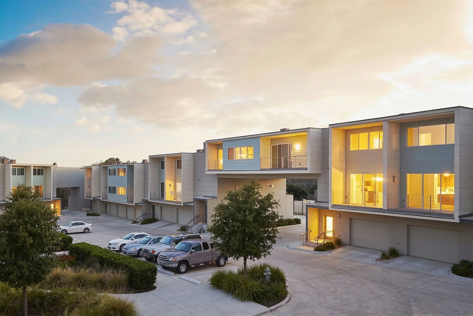 Modern apartment buildings with large glass windows and lit interiors, parked cars in front, landscaped greenery, and a partly cloudy sky at sunset.