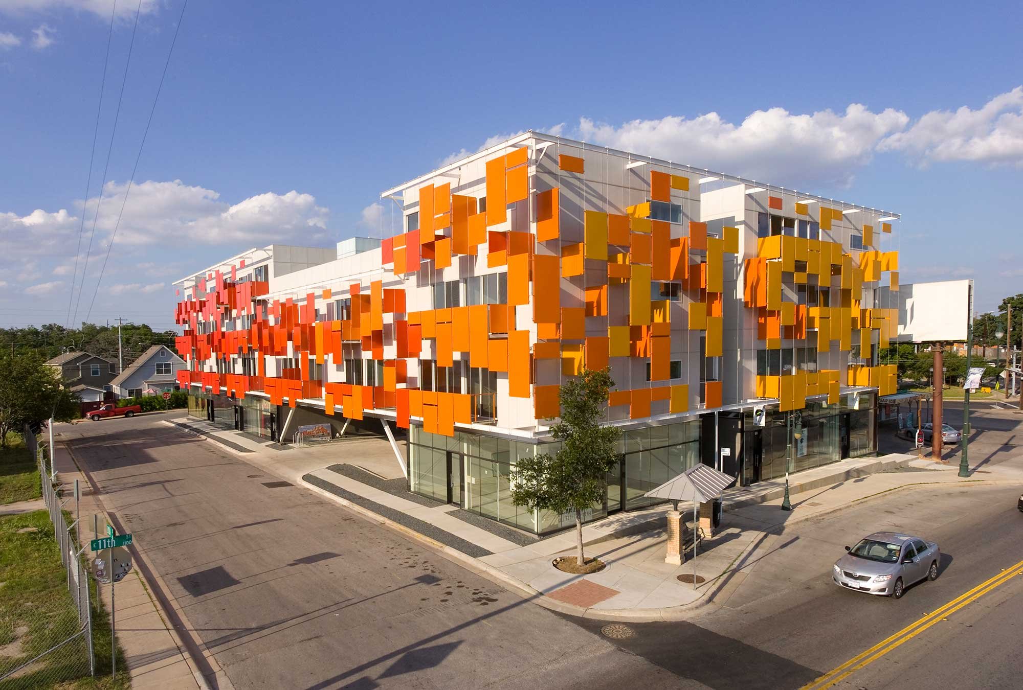 A modern three-story building with a colorful exterior featuring red, orange, and yellow panels, situated on a corner of a city street. The building has large glass windows on the ground floor and is surrounded by a few trees and parked cars.