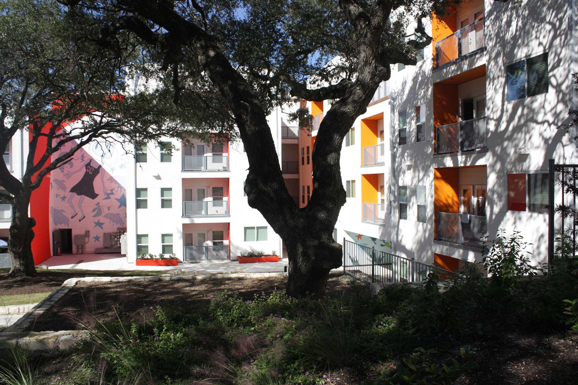 View of a colorful apartment complex with orange and white walls, balconies, and a mural of a dancing figure on one wall. Two large trees with sprawling branches cast shadows over the courtyard area.