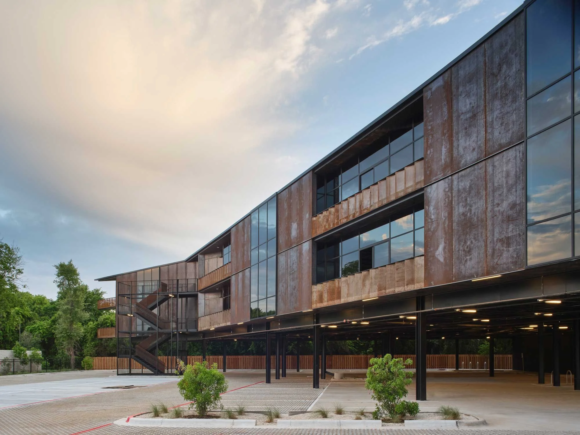 Modern building with rusted metal and glass exterior, outdoor staircase, parking area, and landscaped greenery under a partly cloudy sky.
