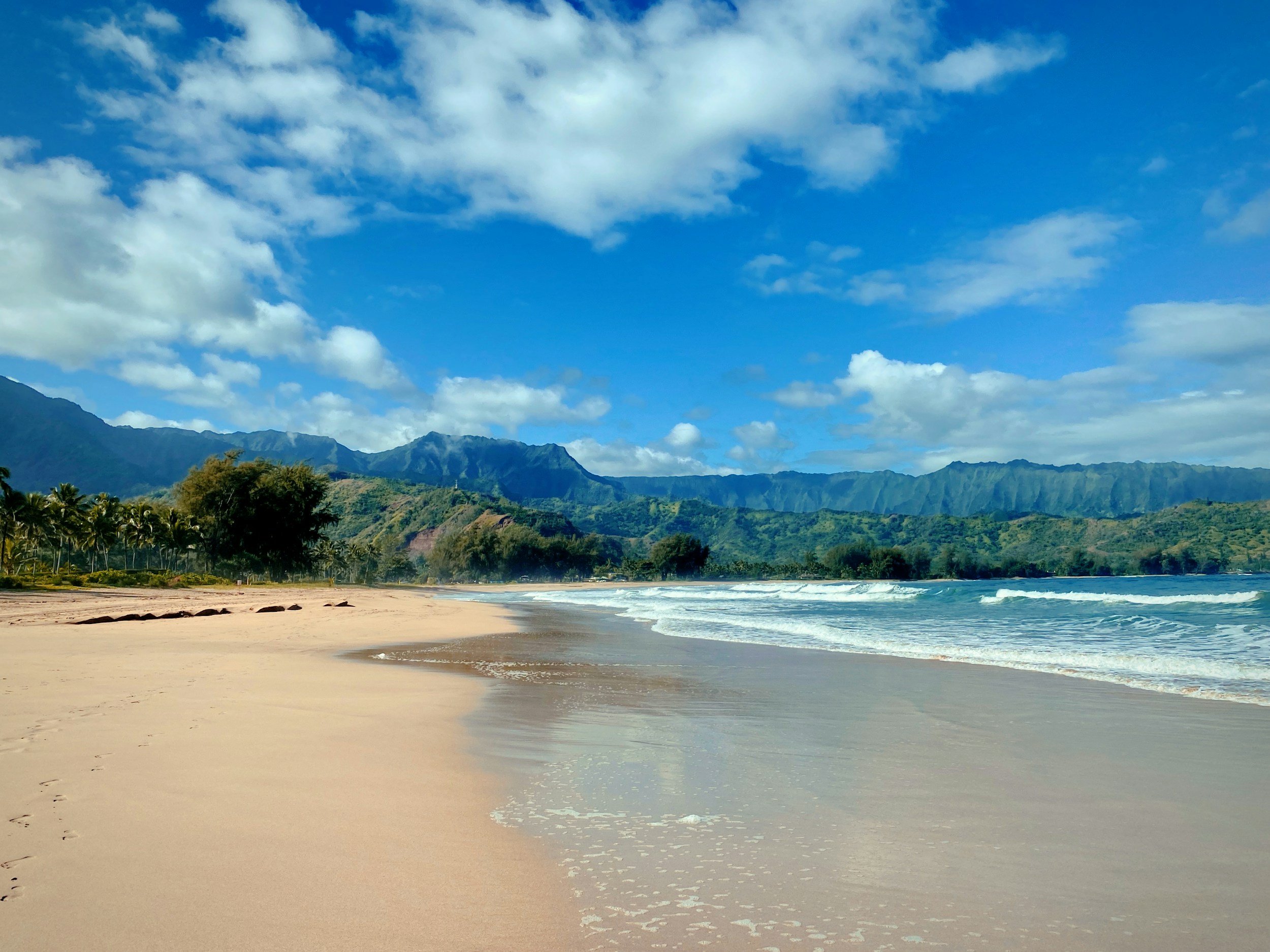 Hanalei Bay beach near Ho’ohiehie ‘Ohana in Princeville, with a sandy shoreline, mountain views, and calm summer water.