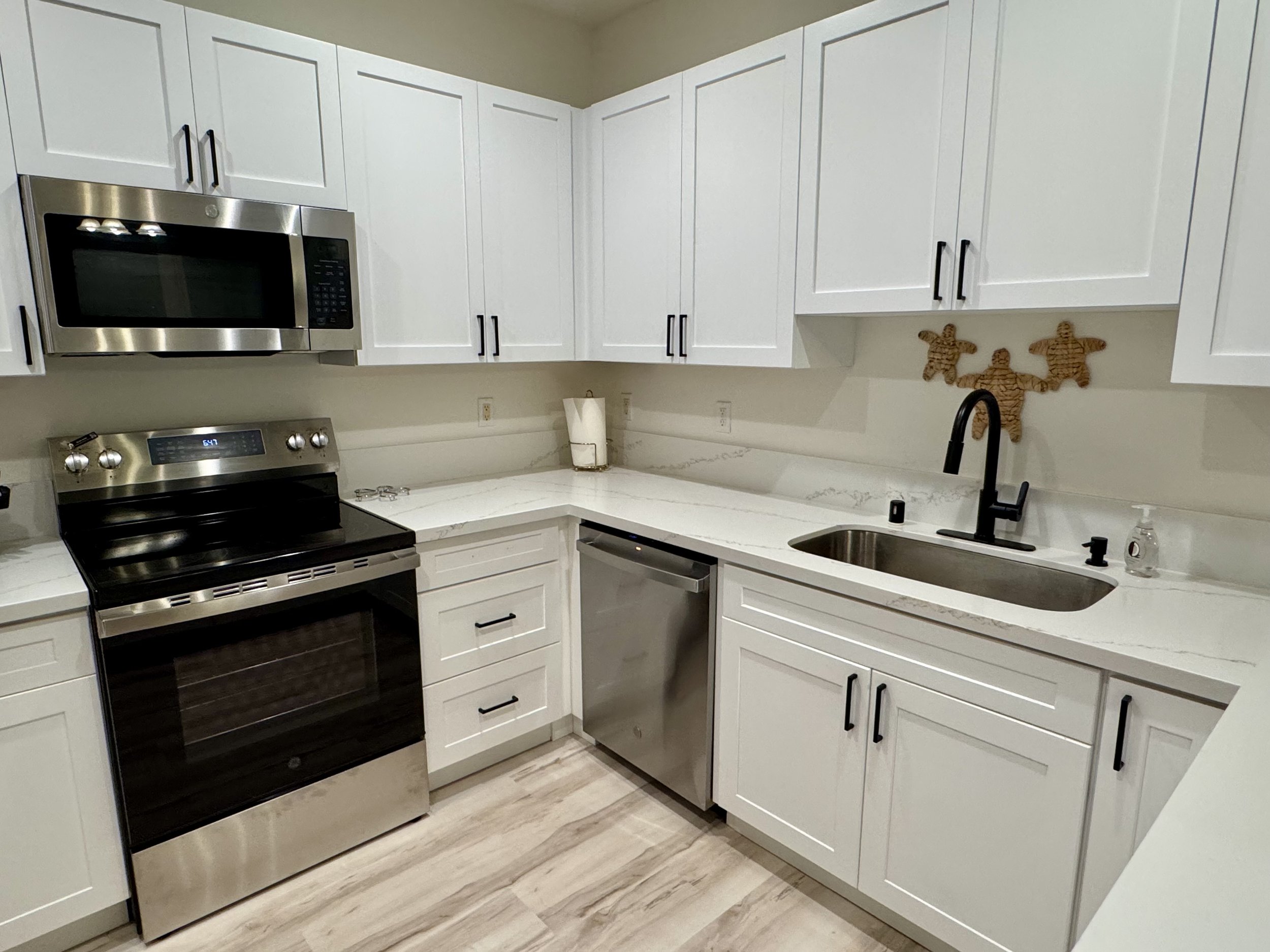 White kitchen with modern appliances, including a microwave above a stove, alongside white cabinets with black handles, a black faucet, a stainless steel dishwasher, and decorated with wicker gingerbread man wall art.