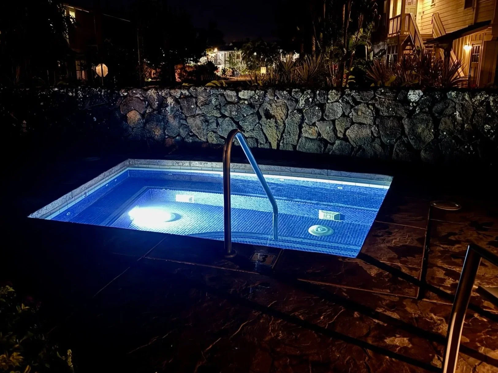 Nighttime scene of a lit-up hot tub or small swimming pool with blue water, stainless steel handrail, surrounded by a stone patio and a stone wall, with houses and plants in the background.