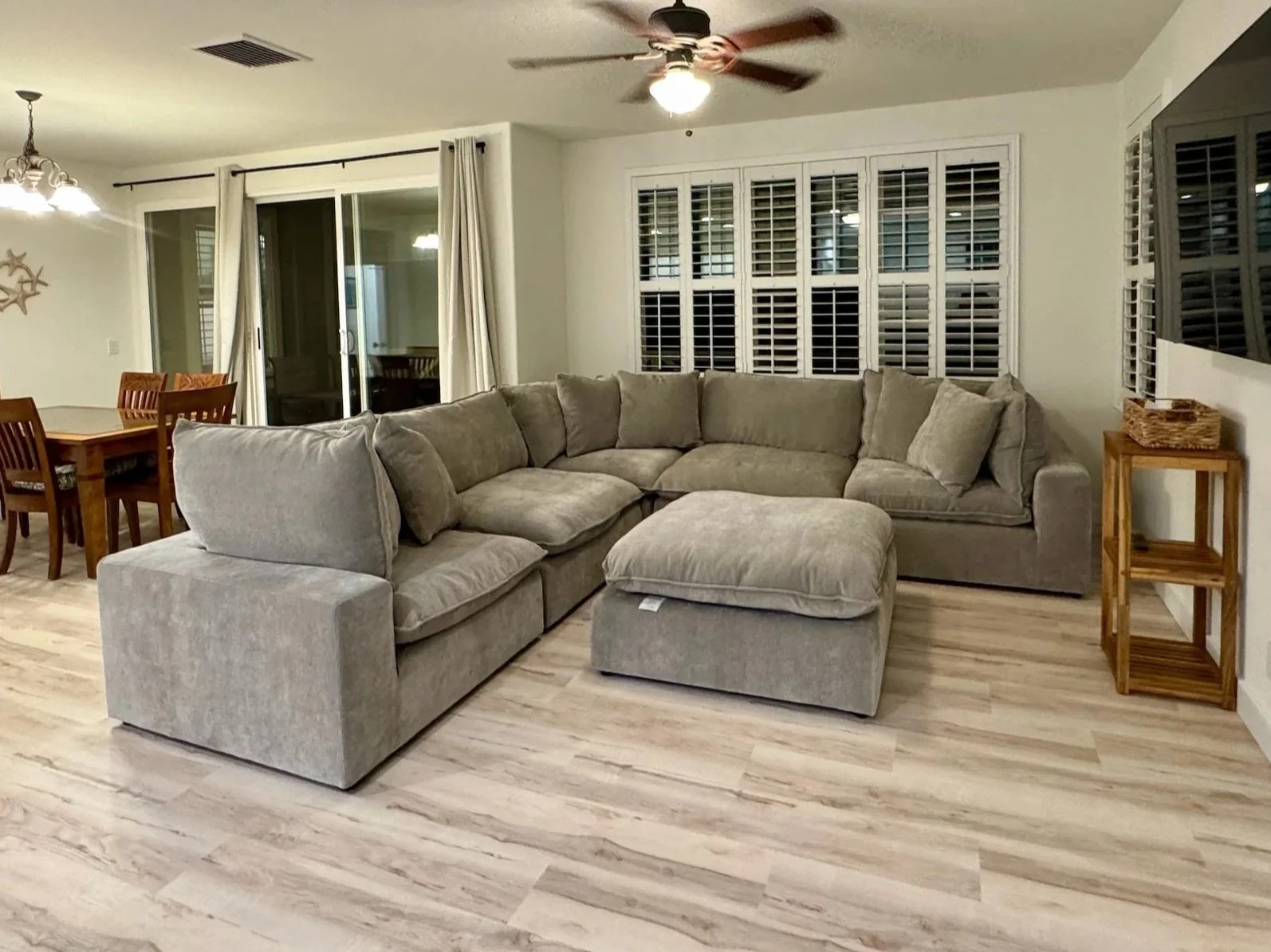The image shows a spacious living room with a large sectional sofa with beige fabric upholstery. Behind the sofa, there are white plantation shutters covering a large window. To the right, a wooden side table with a woven basket on top is visible. To the left, a dining area with a wooden table and matching chairs is partially visible, with sliding glass doors leading to an outdoor space. The room has light-colored wood flooring, a ceiling fan, and a chandelier-style light fixture above the dining area.