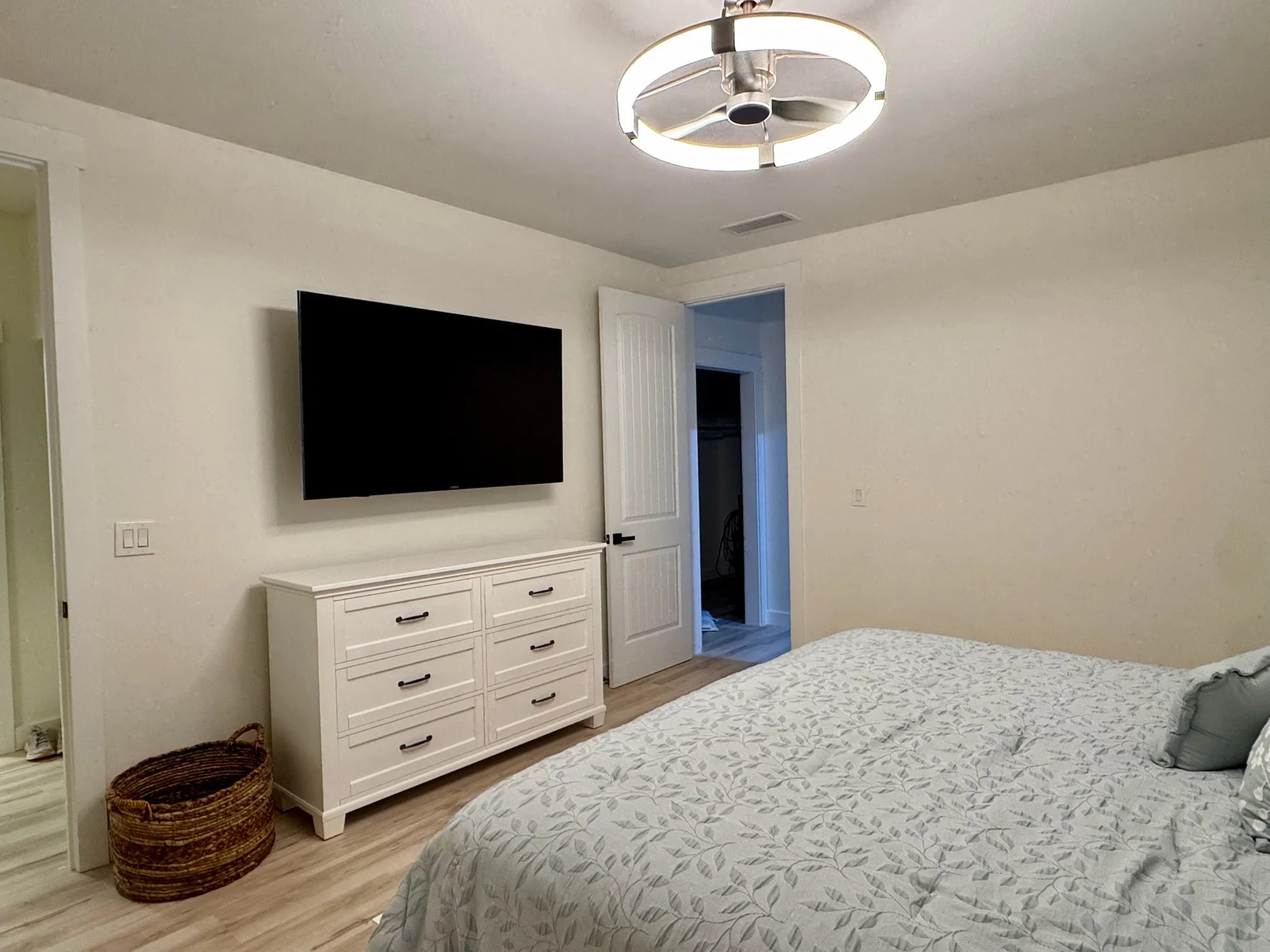 A bedroom with a white dresser, a large flat-screen TV mounted on the wall, a bed with a light-colored patterned comforter, gray pillows, a round woven basket on the floor, and a modern ceiling fan with a light fixture. An open door leads to a closet or hallway.