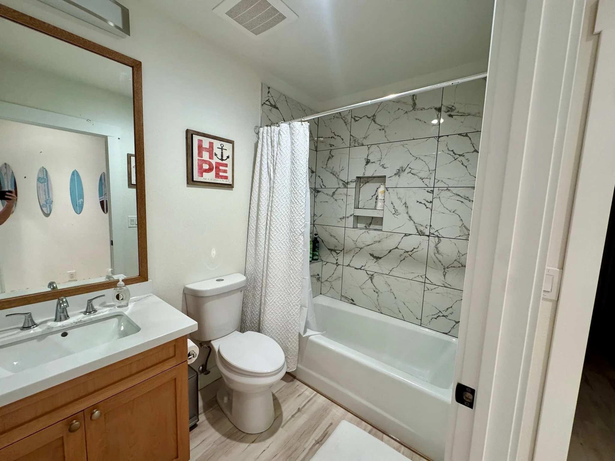 A small bathroom with a shower/tub with marble tile walls, a white shower curtain, a white toilet, a wooden vanity with a sink and faucet, a mirror with a wooden frame, and a wall decoration with the word 'HOPE' and an anchor. There is a white bath mat on the floor.