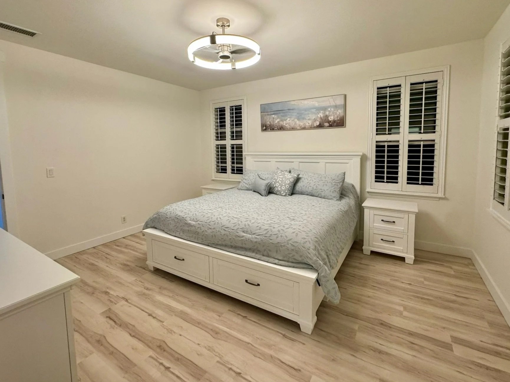 A bedroom with a white bed, gray bedding, and matching pillows, flanked by two white nightstands, and a landscape painting above the headboard, with three windows with white shutters and light wood flooring.