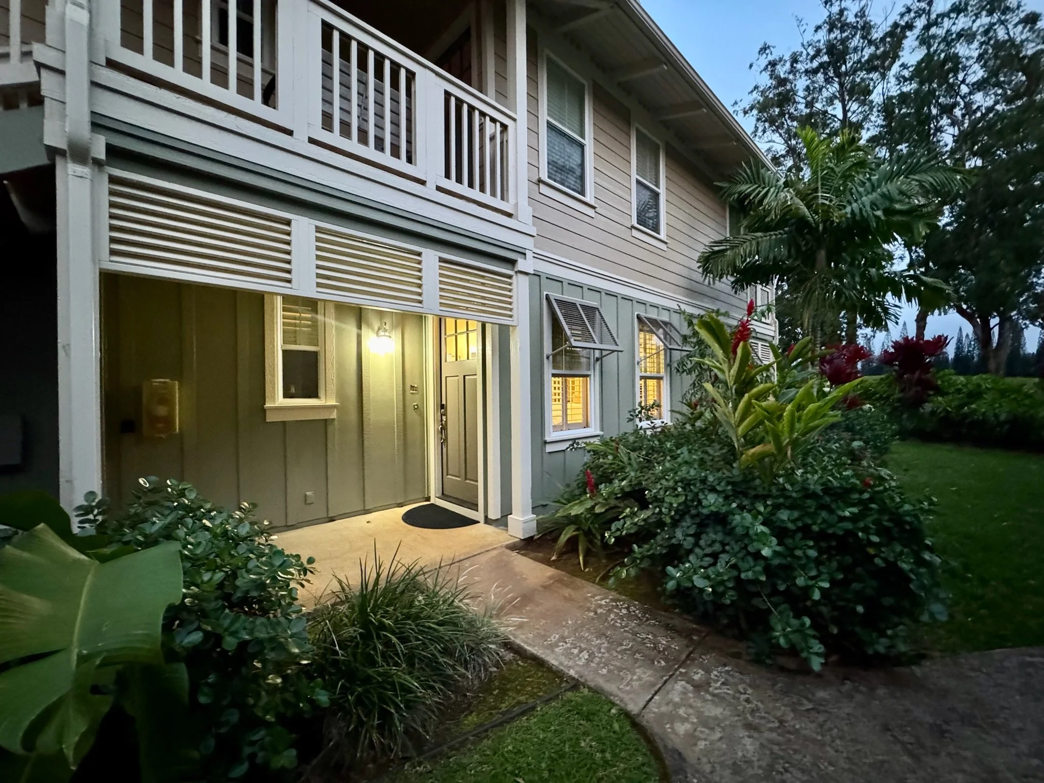 The front entrance of a house at dusk, with illuminated windows, a door light, a pathway, and surrounding greenery including bushes and a mini palm tree.