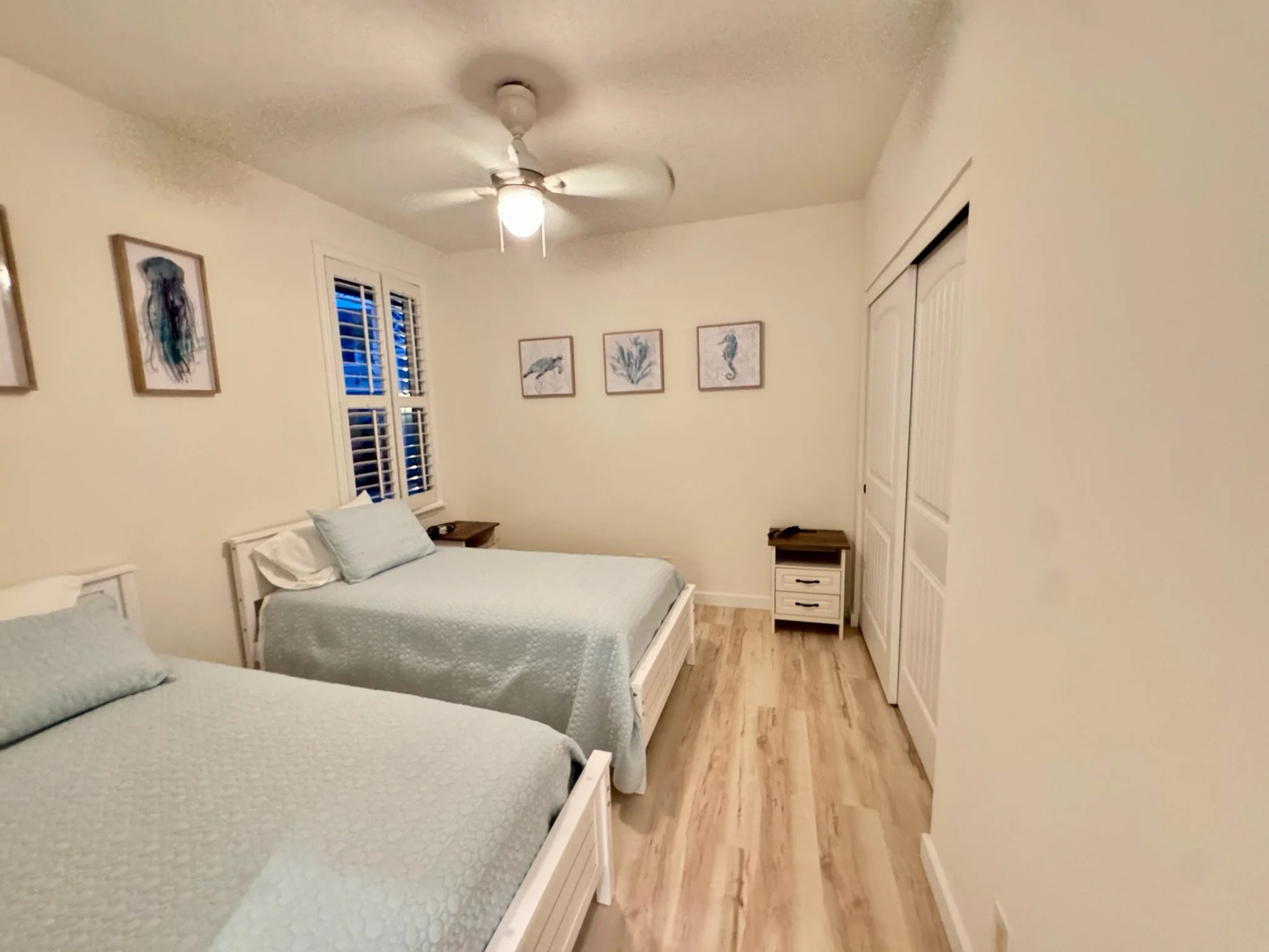 A bedroom with two beds, one in the foreground and one towards the back, each with light-colored bedding and pillows. The room has wooden flooring, a ceiling fan with a light, and a window with shutters on the left wall. The walls are decorated with framed pictures of sea creatures and plants. There are nightstands beside each bed, and a closet with sliding doors on the right.