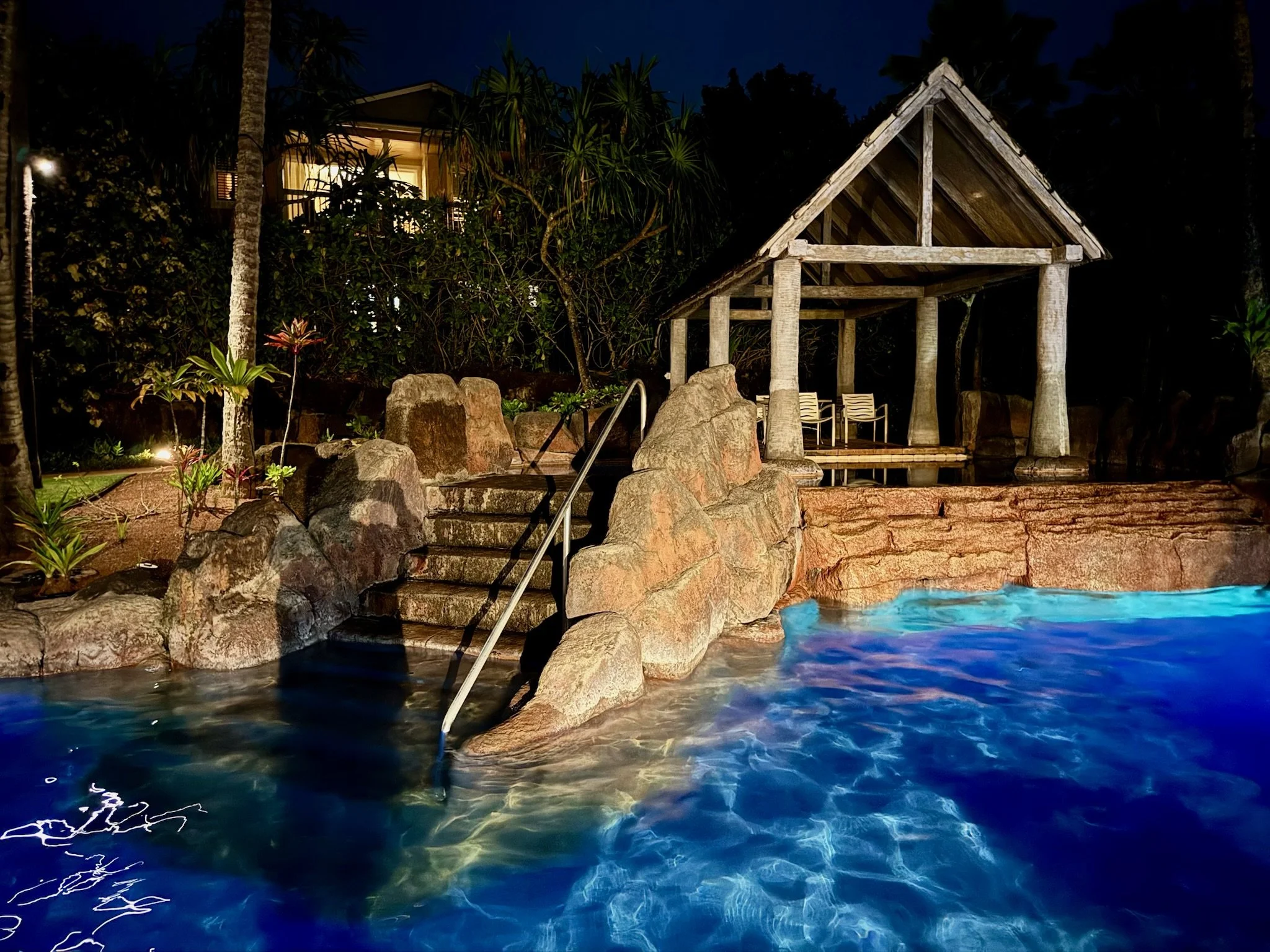 Nighttime view of a resort pool area with stone steps leading into the water, a wooden gazebo with chairs, lush tropical plants, and a lit building in the background.
