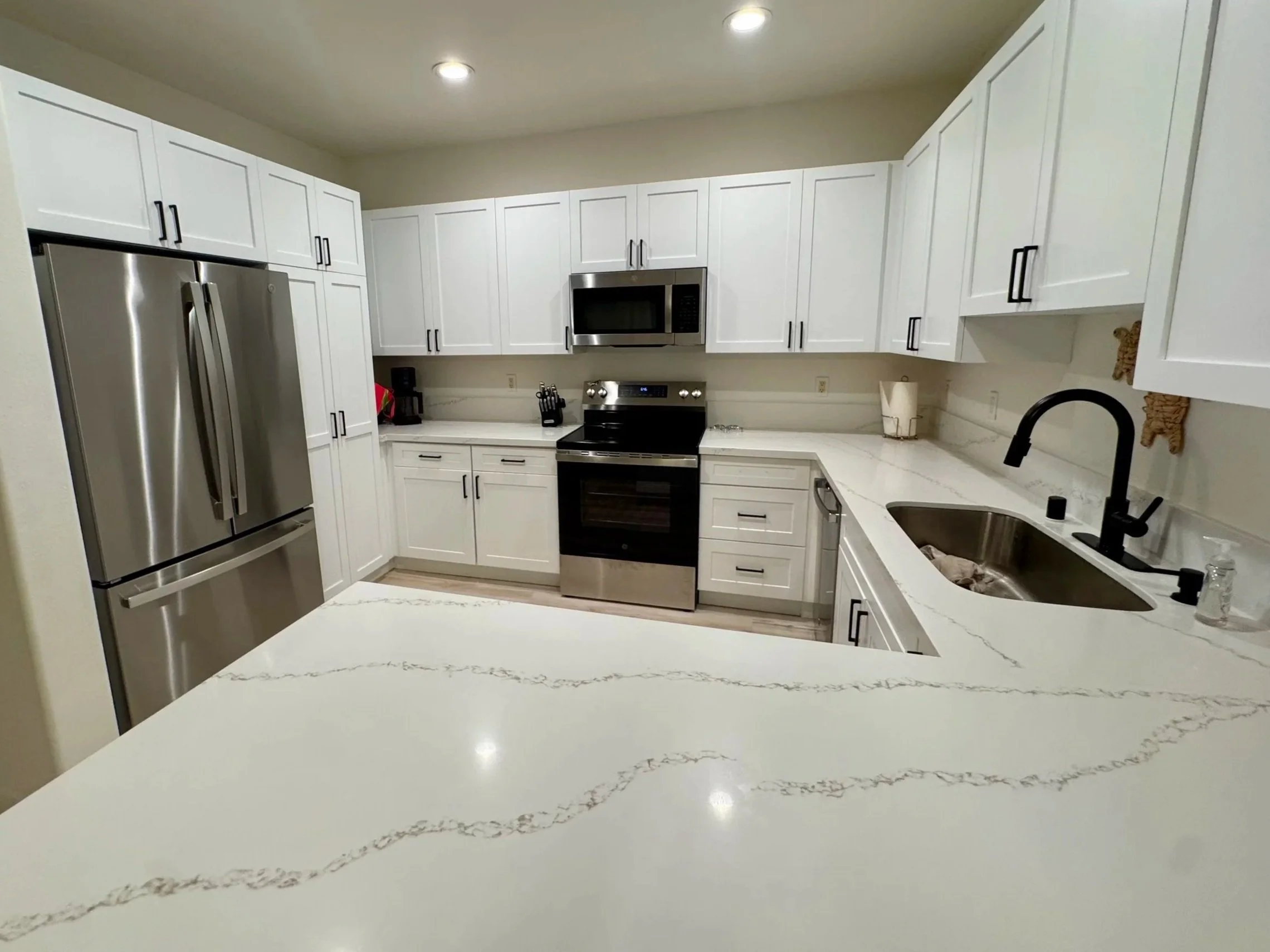 Modern kitchen with white cabinets, stainless steel refrigerator, oven, microwave, and black faucet, with a marble countertop and beige walls.