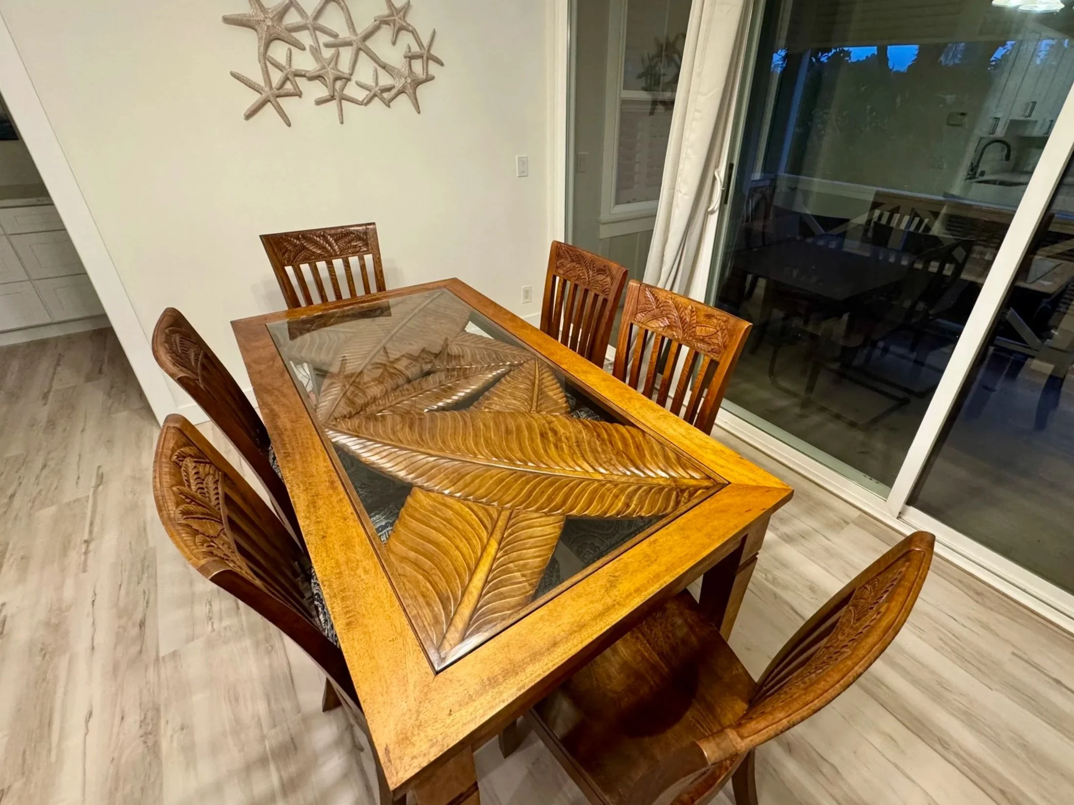 Wooden dining table with a glass top featuring carved leaves, surrounded by six matching wooden chairs with carved leaf patterns on the backrests, located in a dining room with a sliding glass door and a wall decoration of starfish.