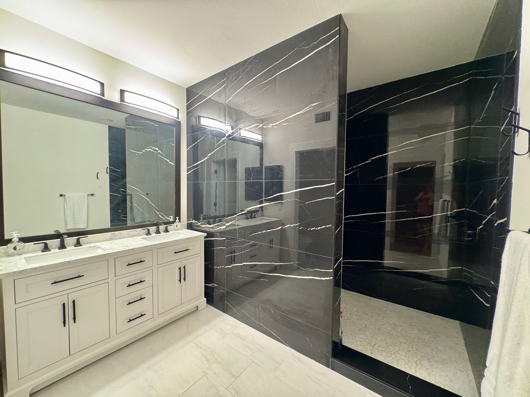 Modern bathroom with white vanity, black faucet, large mirror, black marble walk-in shower with a pebble floor.