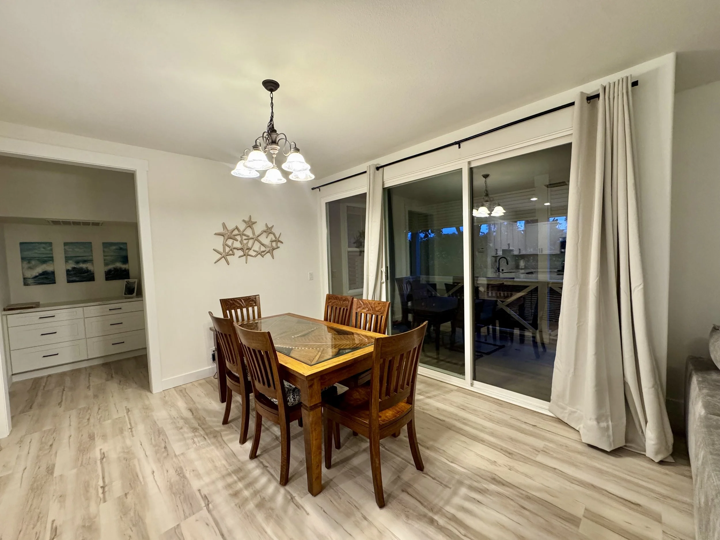 Dining room with a wooden table and six matching chairs, sliding glass doors with curtains, and a chandelier light fixture.