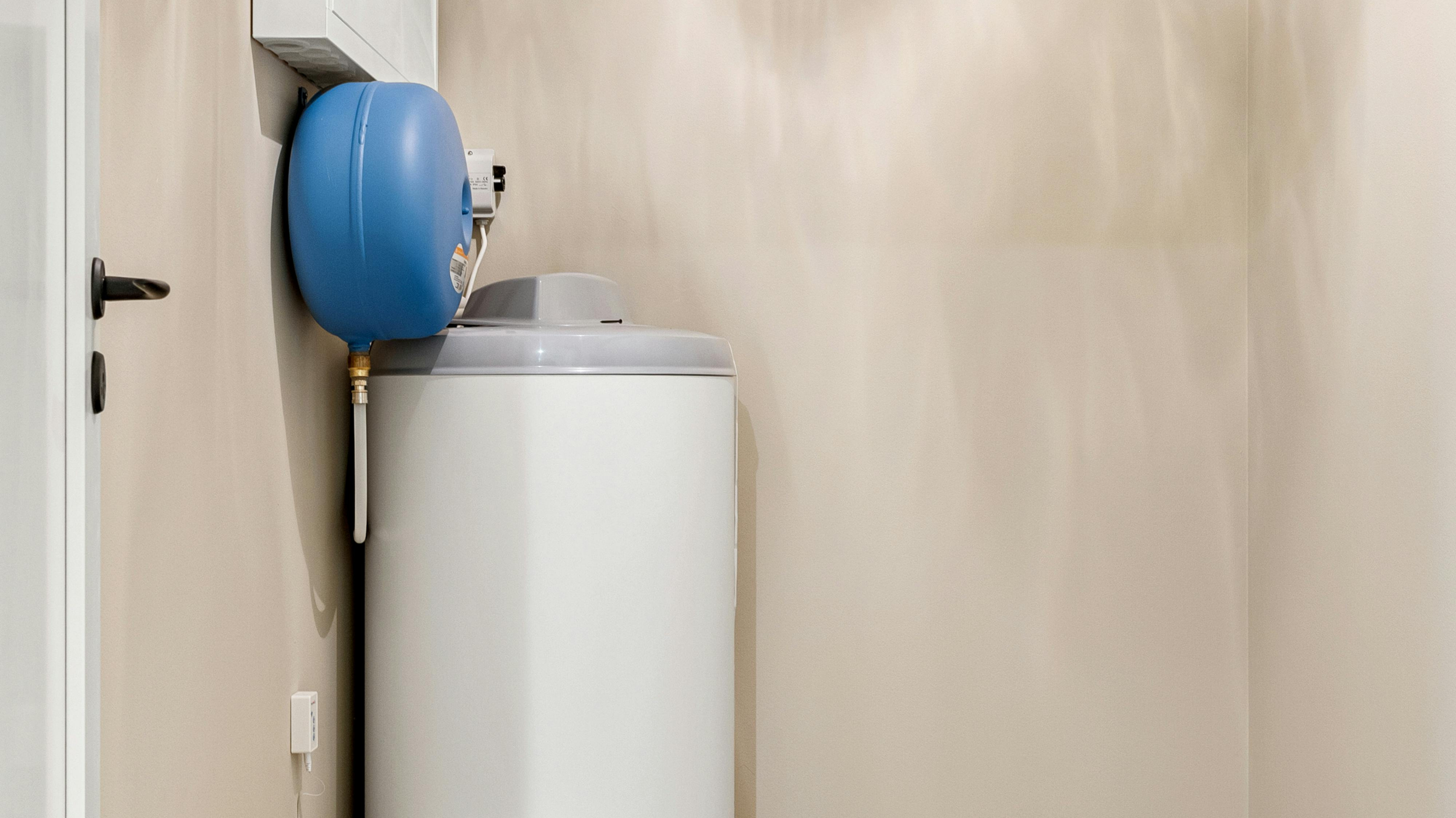 A laundry room with a top-loading washing machine and a blue water pressure tank on top, set against beige walls.