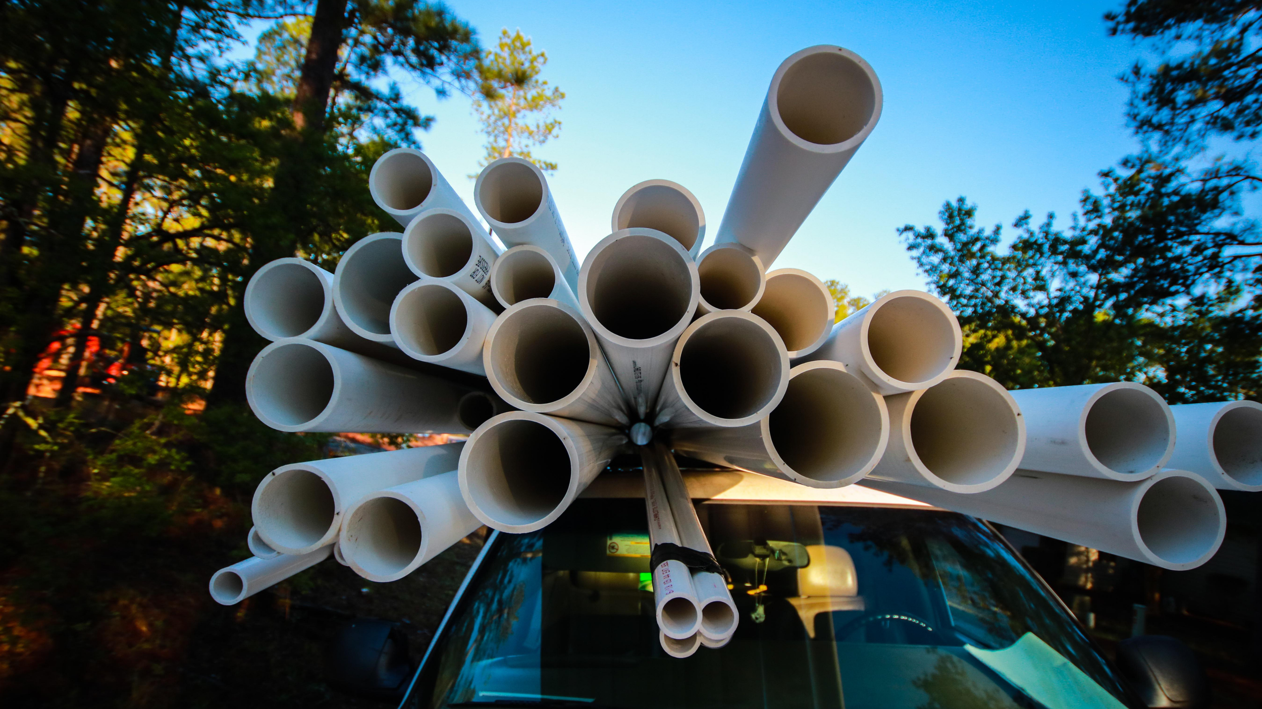 A vehicle with a large bundle of white PVC pipes on its roof, set against a background of trees and blue sky.