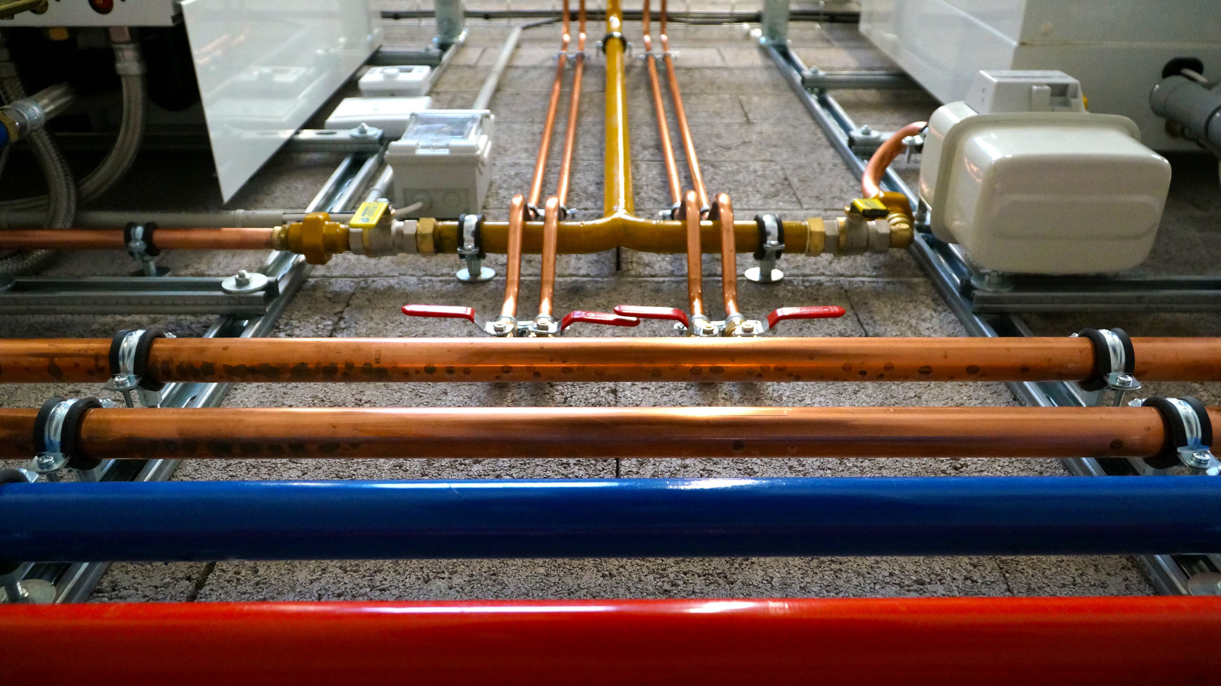 A mechanical room with copper and blue pipes, valves, and electrical conduits installed on a concrete floor.