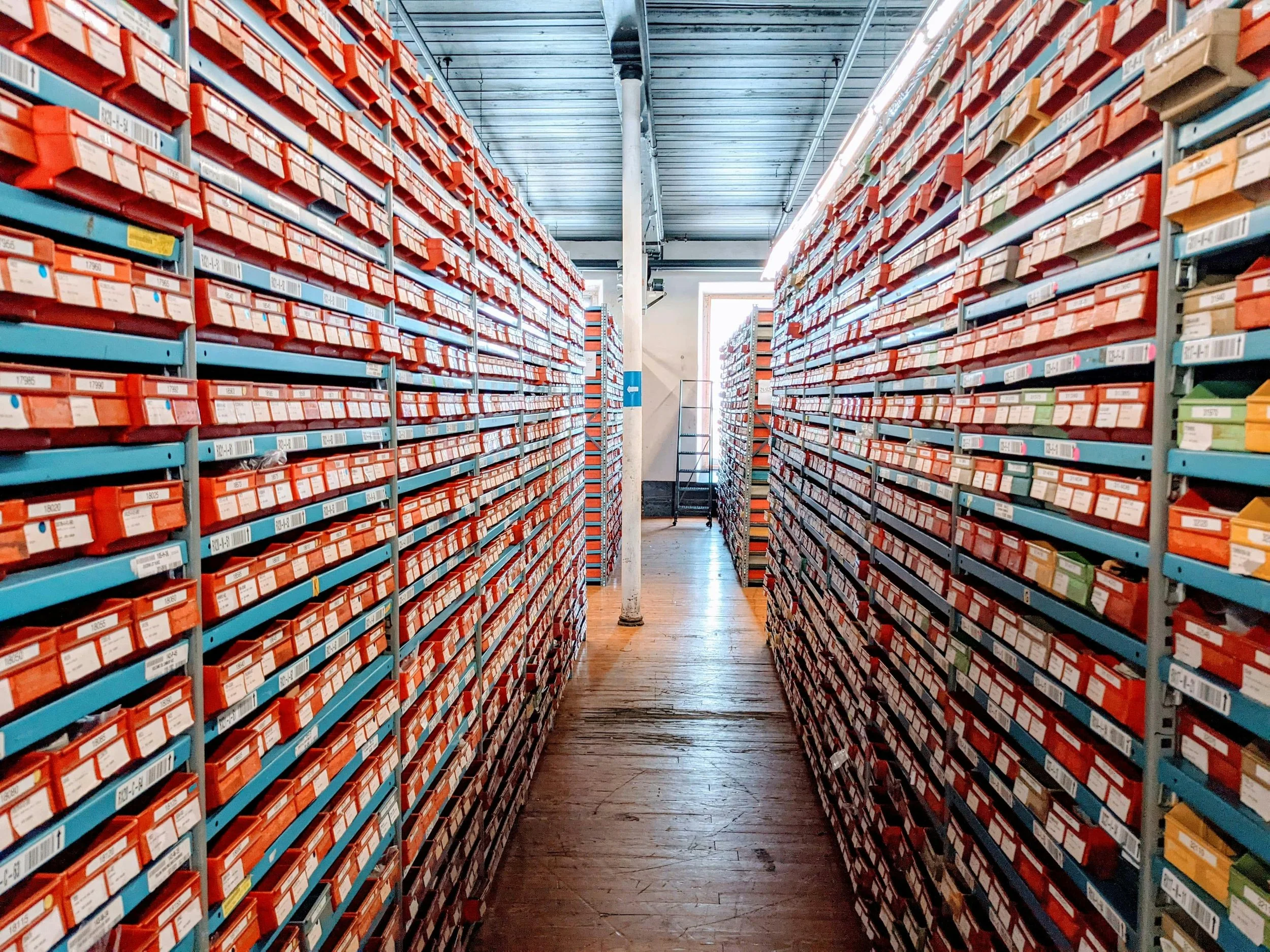 A storage room with tall metal shelves filled with small labeled boxes, creating a narrow aisle between them, with a window at the end of the aisle.