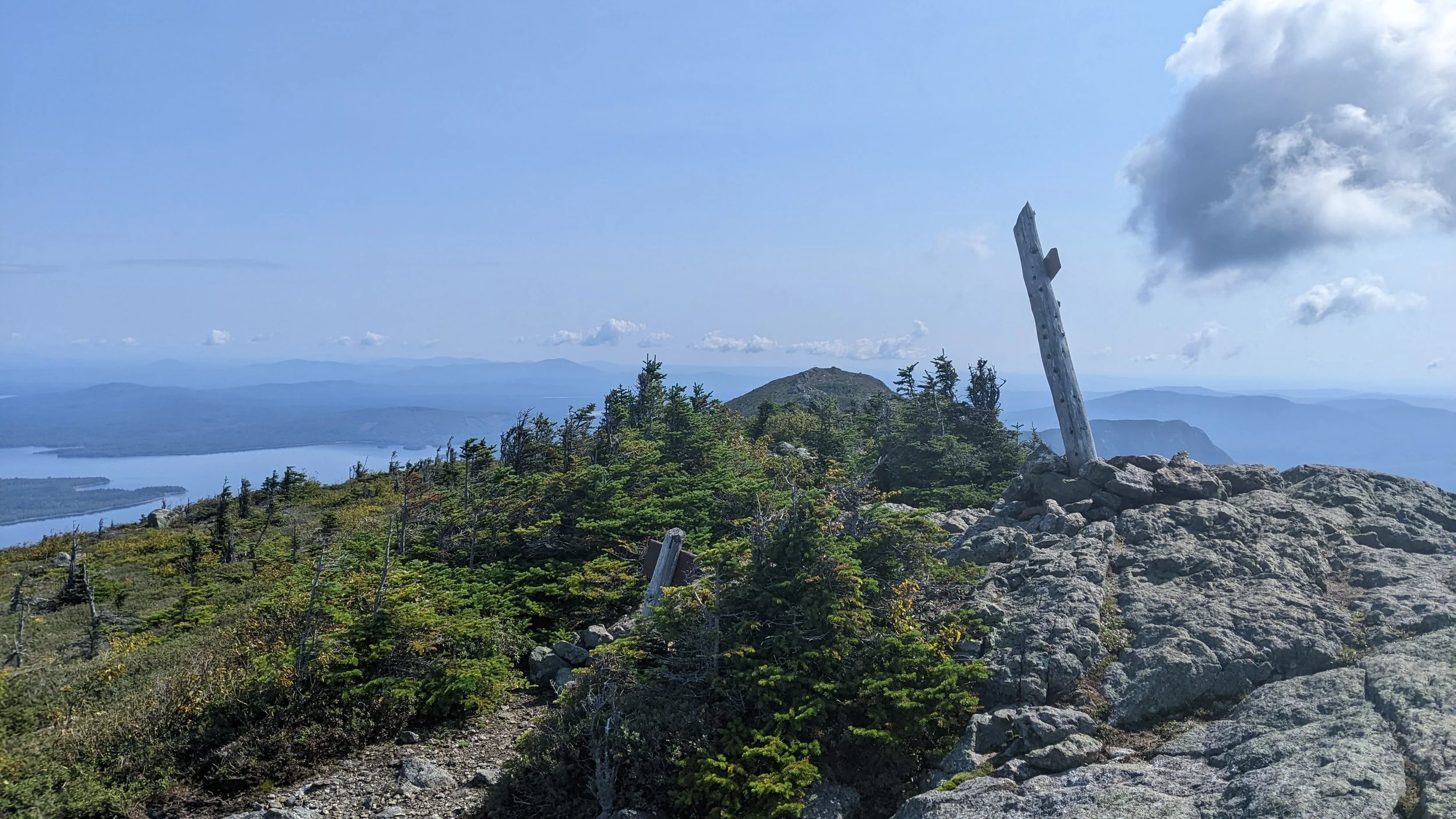 Bigelow Range, Maine. Mountain summit with a wooden post leaning on rocks, surrounded by green shrubs and trees, with a lake and distant mountains under a partly cloudy sky.
