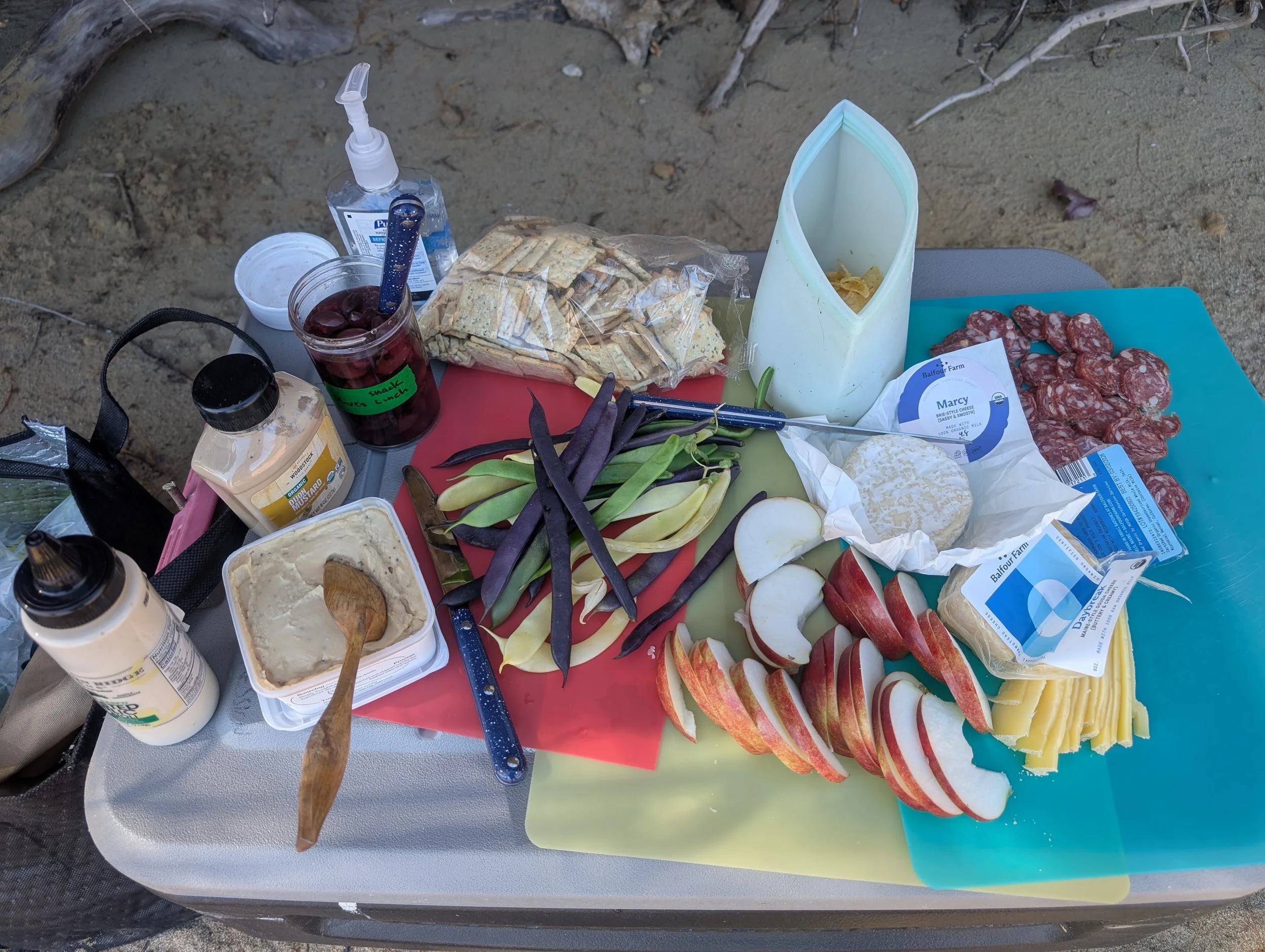 A picnic table with various foods including sliced apples, cheese, cured meats, crackers, chips, and a jar of cherries, set outdoors on sandy ground.