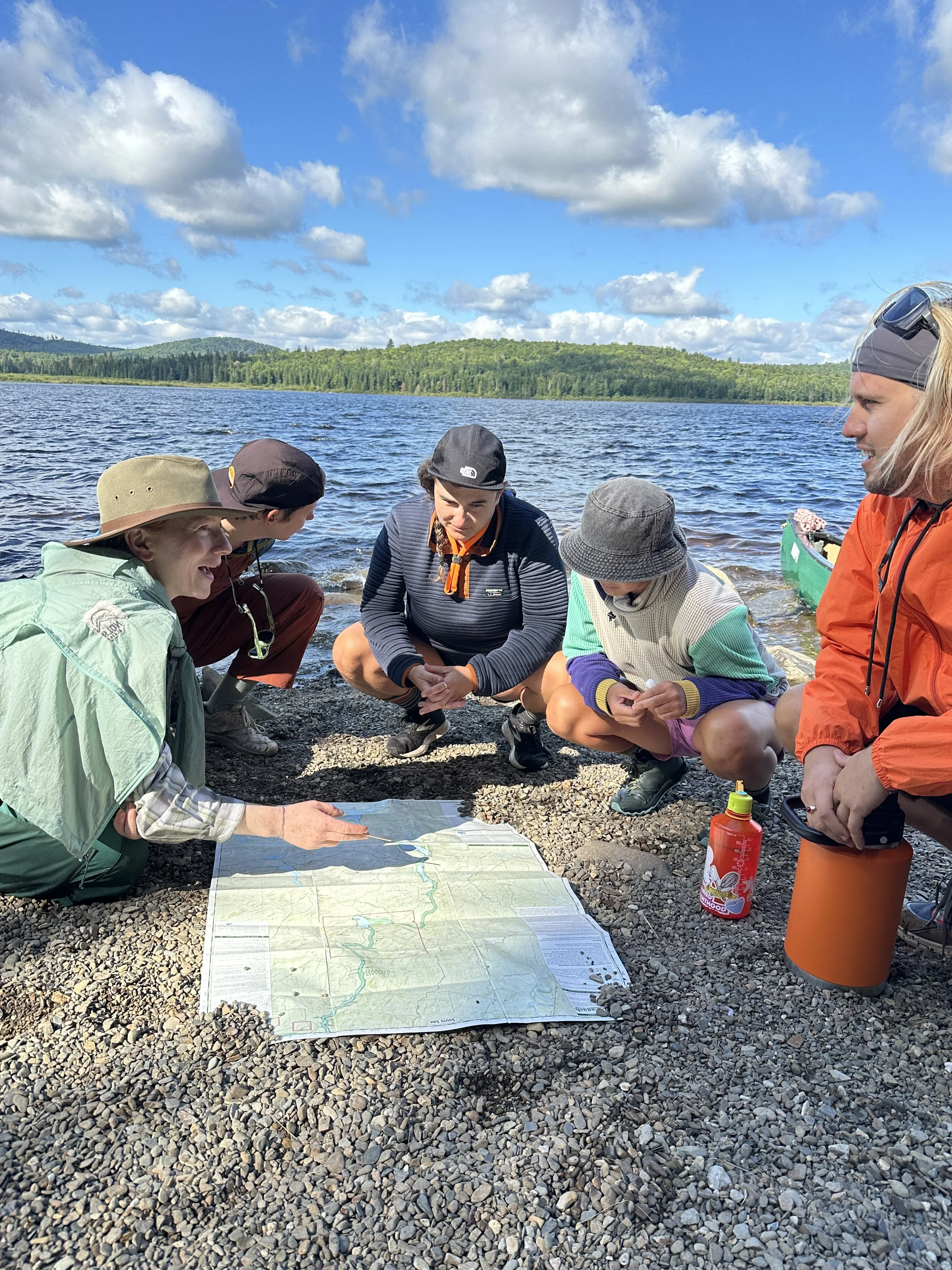 A group of five people sitting and kneeling on a rocky shoreline by a lake, examining a large map spread on the ground. They are outdoors under a partly cloudy sky with green hills and trees in the background. One person has a bottle of water nearby 