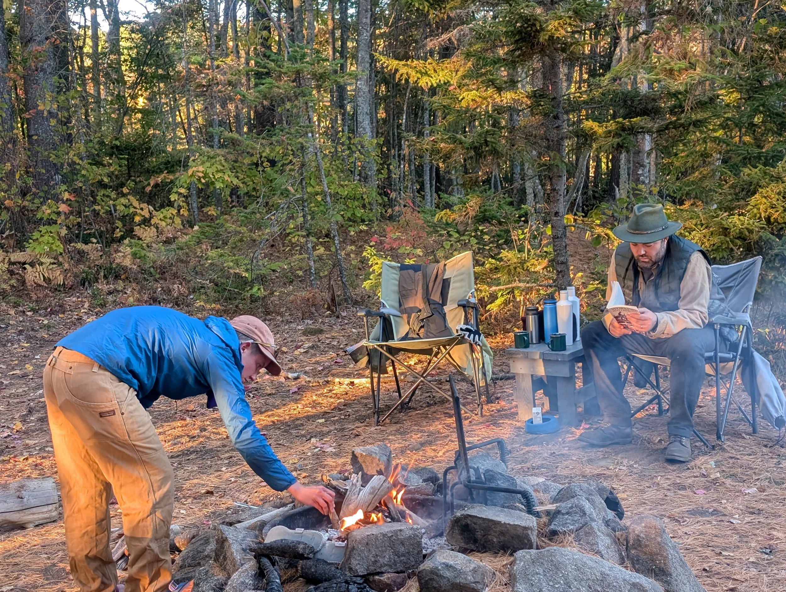 Two people camping outdoors in a forested area during sunset. One person is kneeling near a campfire, and the other is sitting on a camping chair reading a book. Camping gear and chairs are set up around them, with trees and natural scenery in the ba
