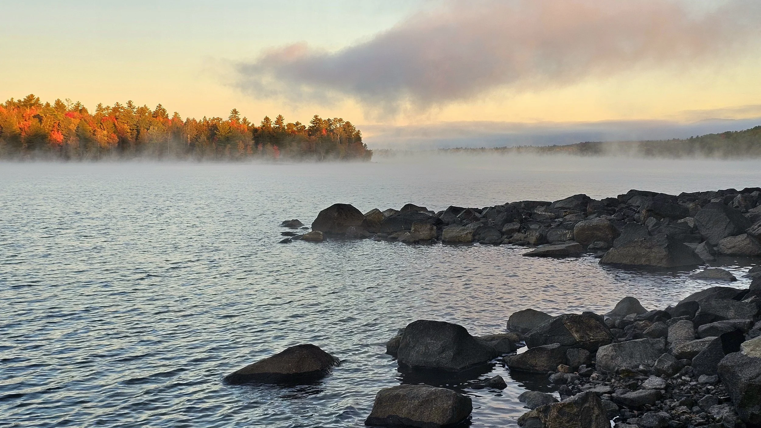 Flagstaff Lake, Maine. A lakeside scene at sunrise with calm water, a rocky shoreline in the foreground, a mist over the water, and a forest with autumn-colored trees in the background, under a partly cloudy sky.
