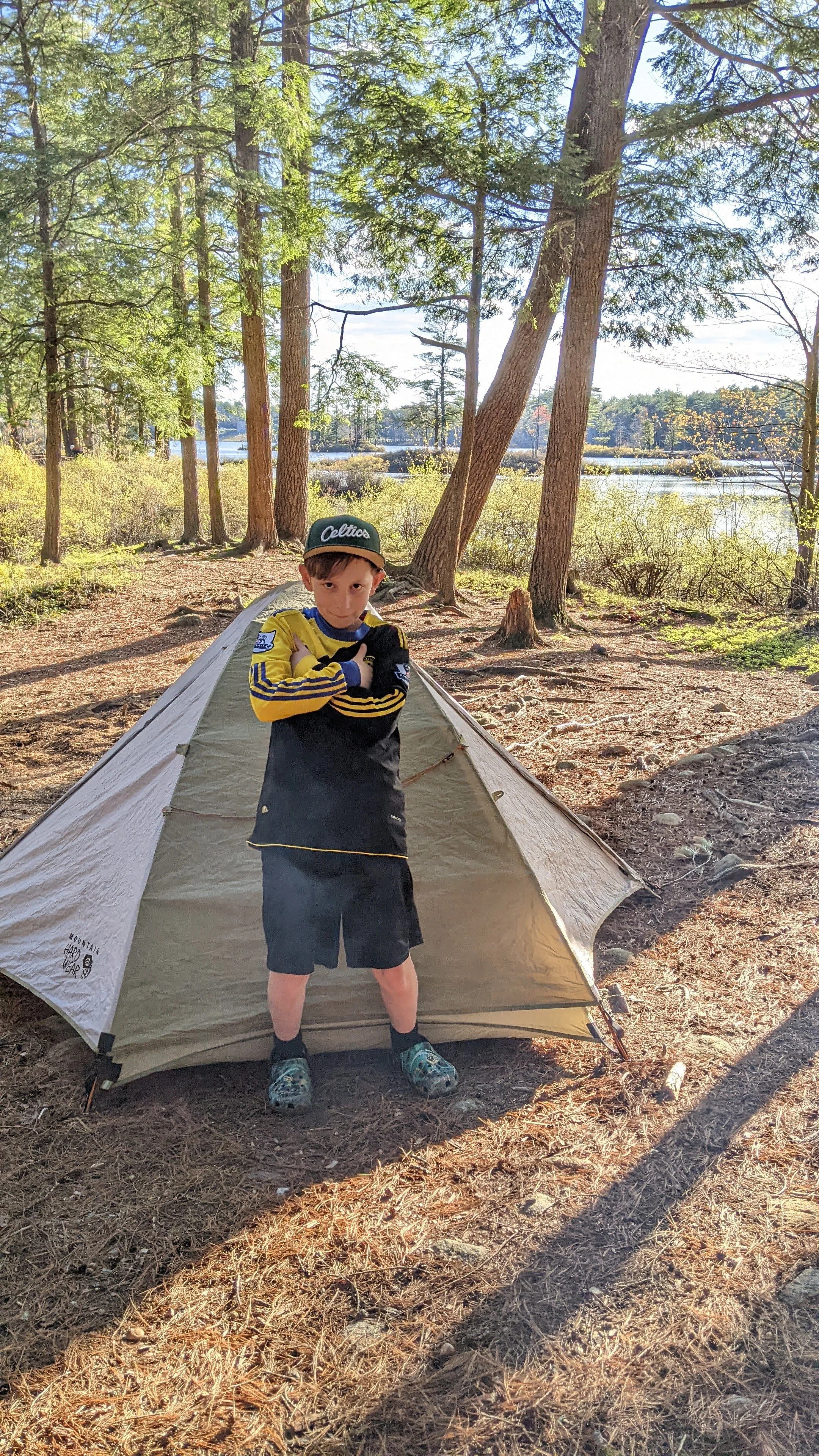 A young boy wearing a cap, a sports jersey, shorts, and crocs stands in front of a camping tent in a wooded area near a lake, with trees and water in the background.
