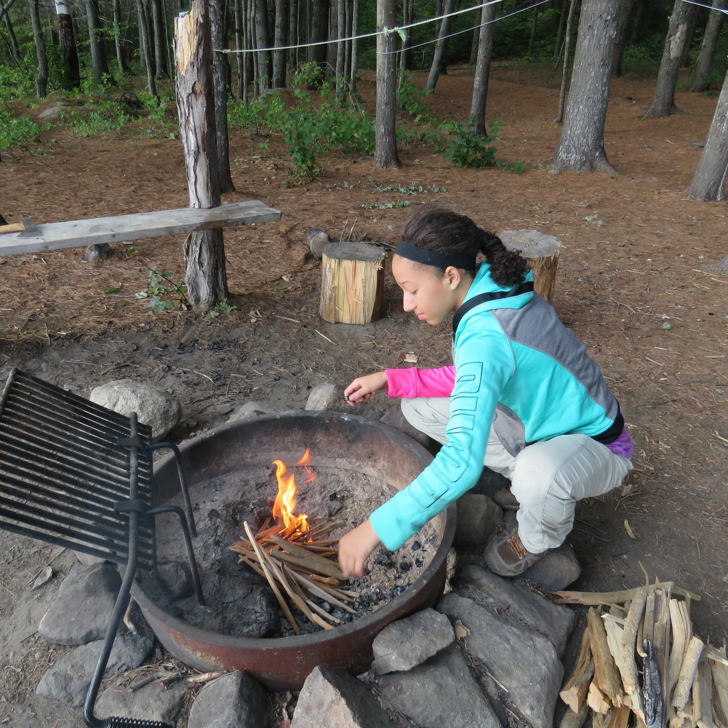 A person with curly hair wearing a black headband and a blue hoodie crouching near an outdoor campfire grill in a wooded area. The campfire is in a round metal fire pit with logs and sticks, and small flames are visible. In the background, there are 