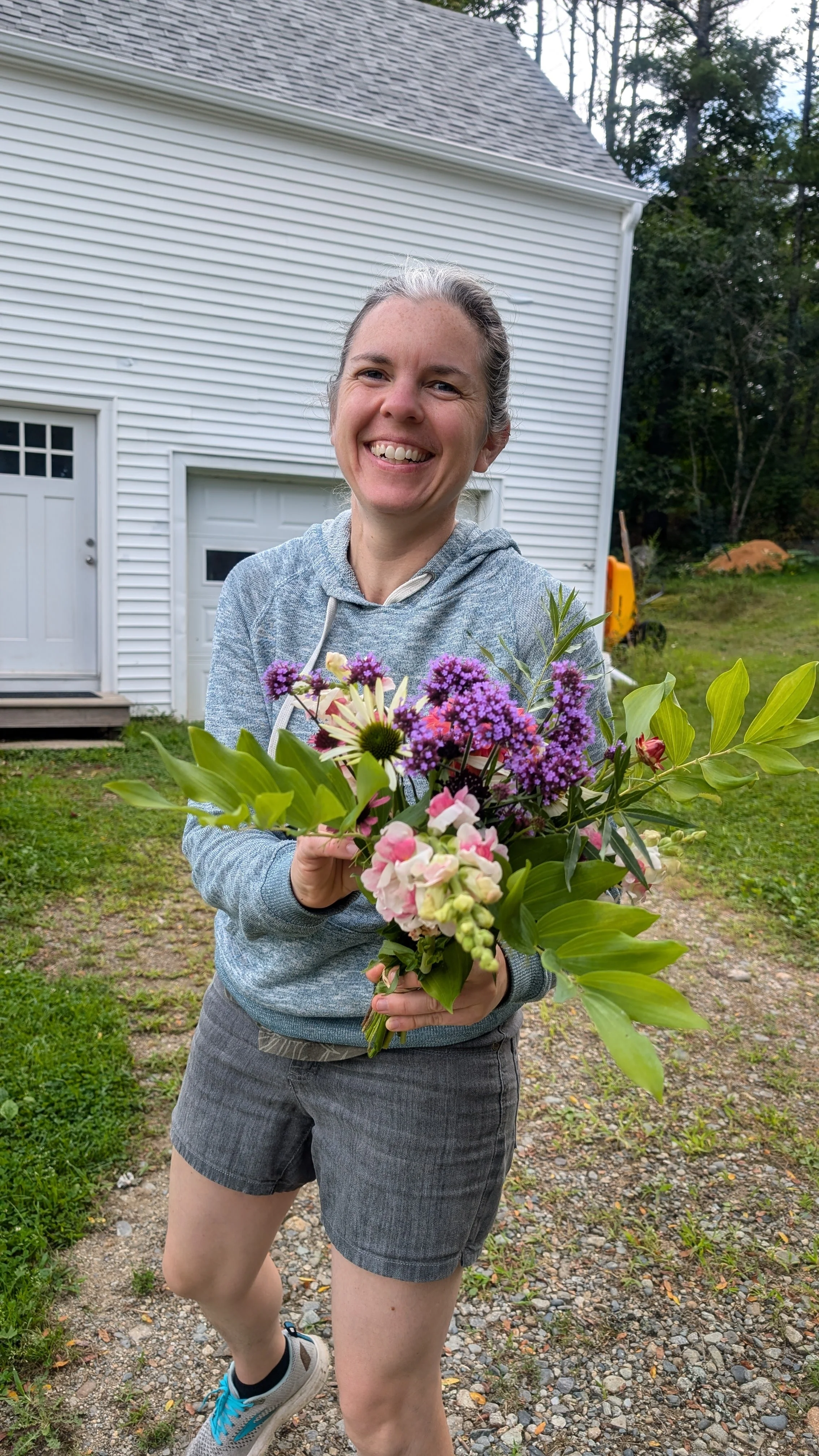 Flowers from the garden. A woman smiling and holding a bouquet of colorful flowers outside in front of a white house garage with trees in the background.