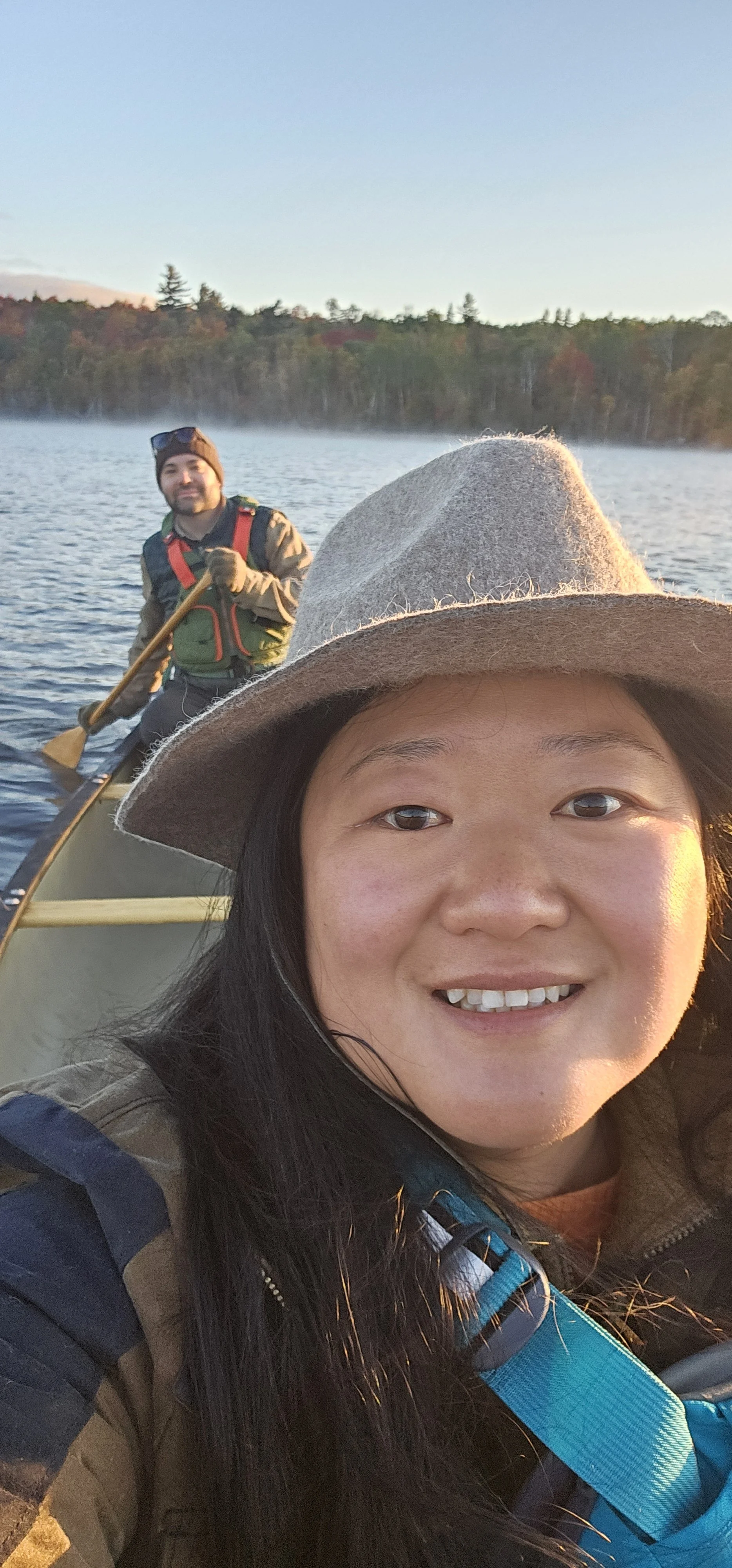 A woman taking a selfie on a boat with a man paddling in the background on a lake during sunset.