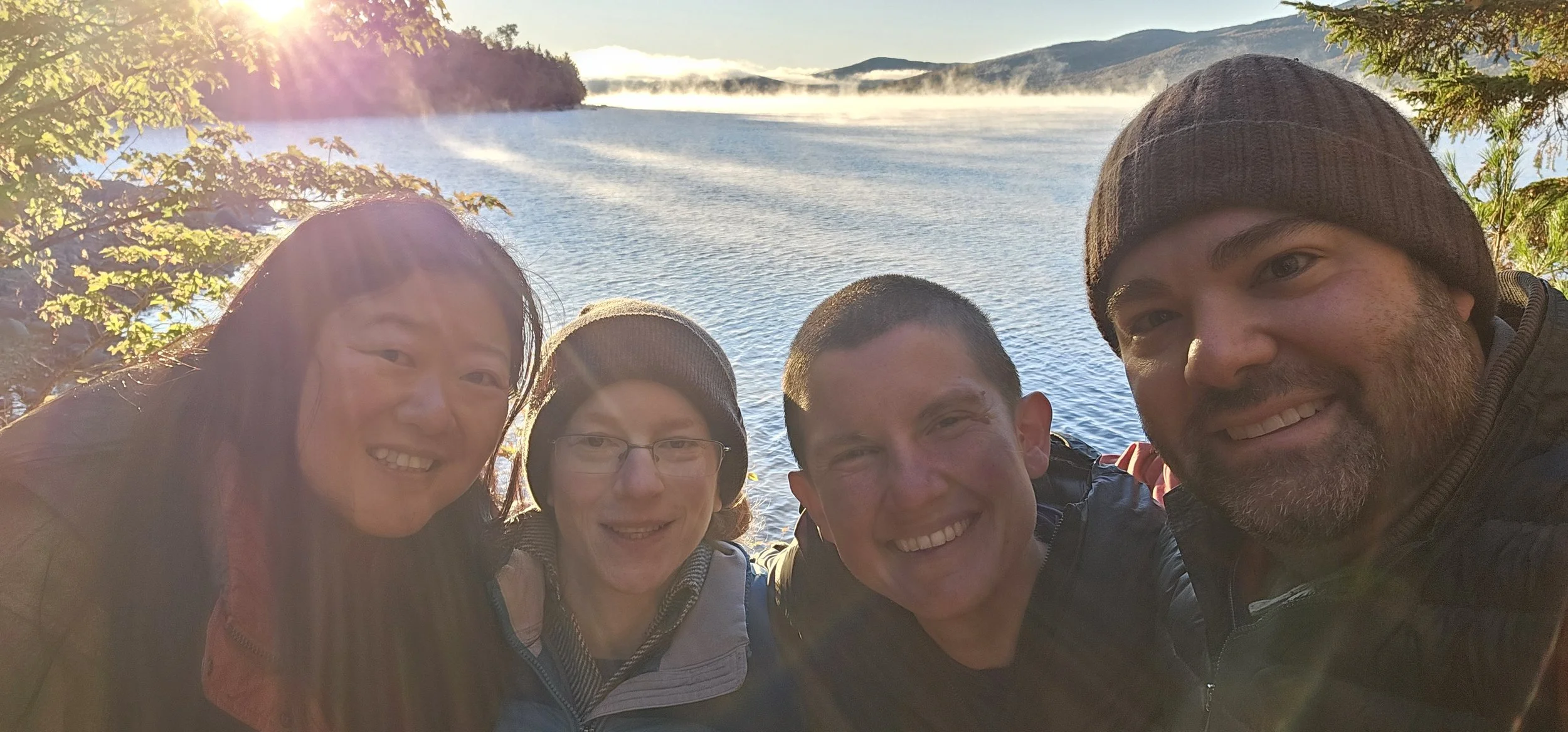 Group of five people taking a selfie near a lake with hills and mist in the background, during sunrise or sunset.