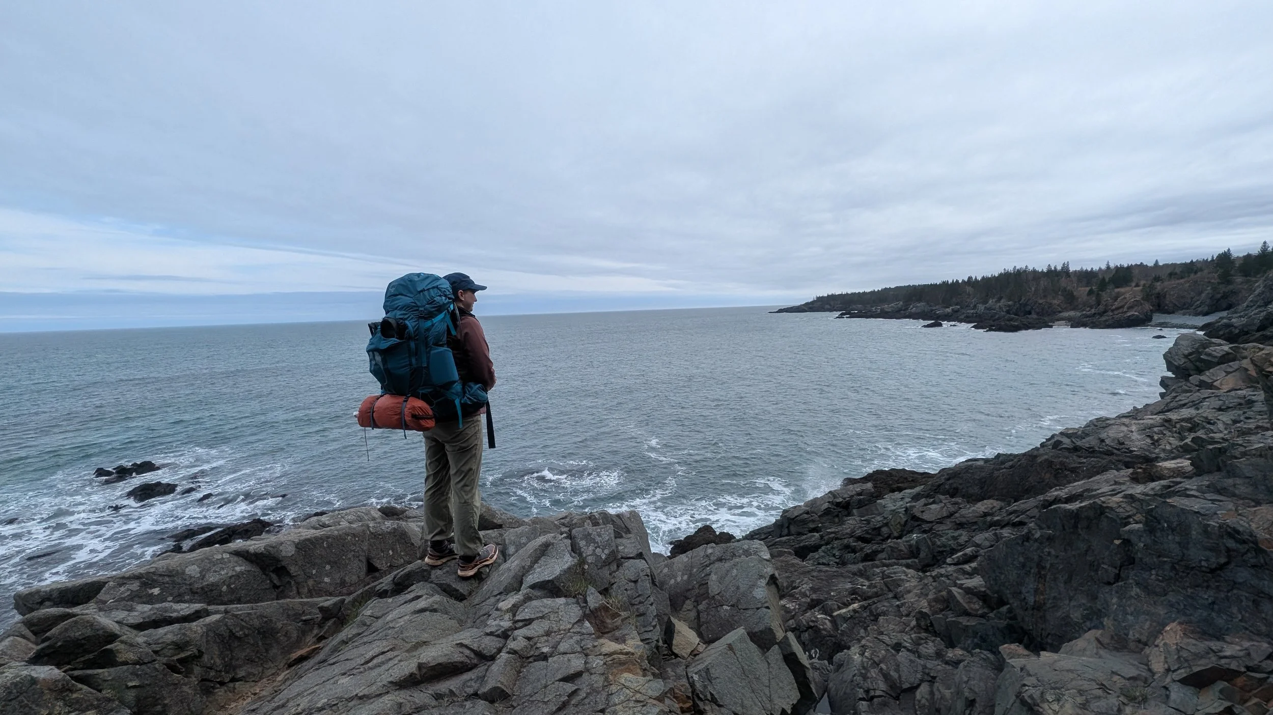 Bold Coast Trail Cutler, Maine. A person with a backpack standing on rocky shoreline overlooking the ocean and distant cliffs under cloudy sky.