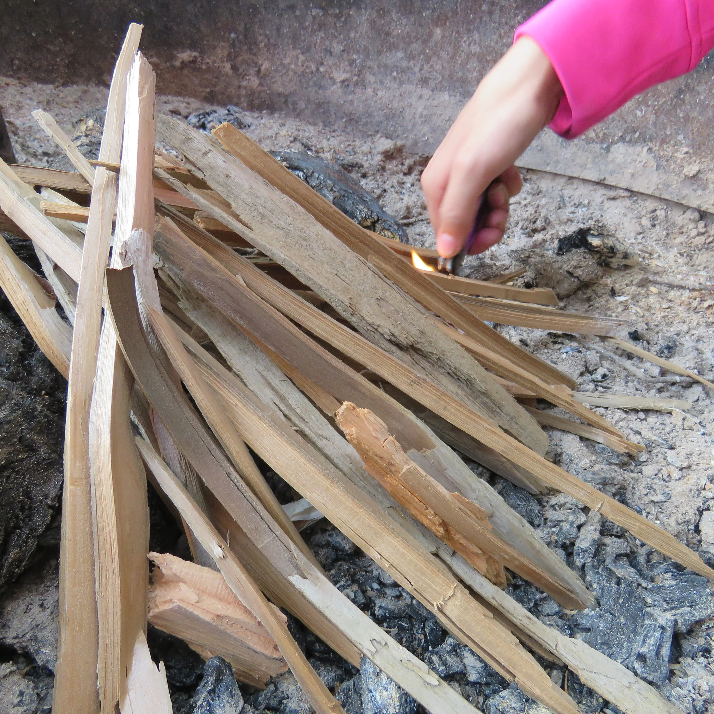 A hand holding a matchstick igniting a small fire on wooden sticks arranged for a campfire on rocky ground.