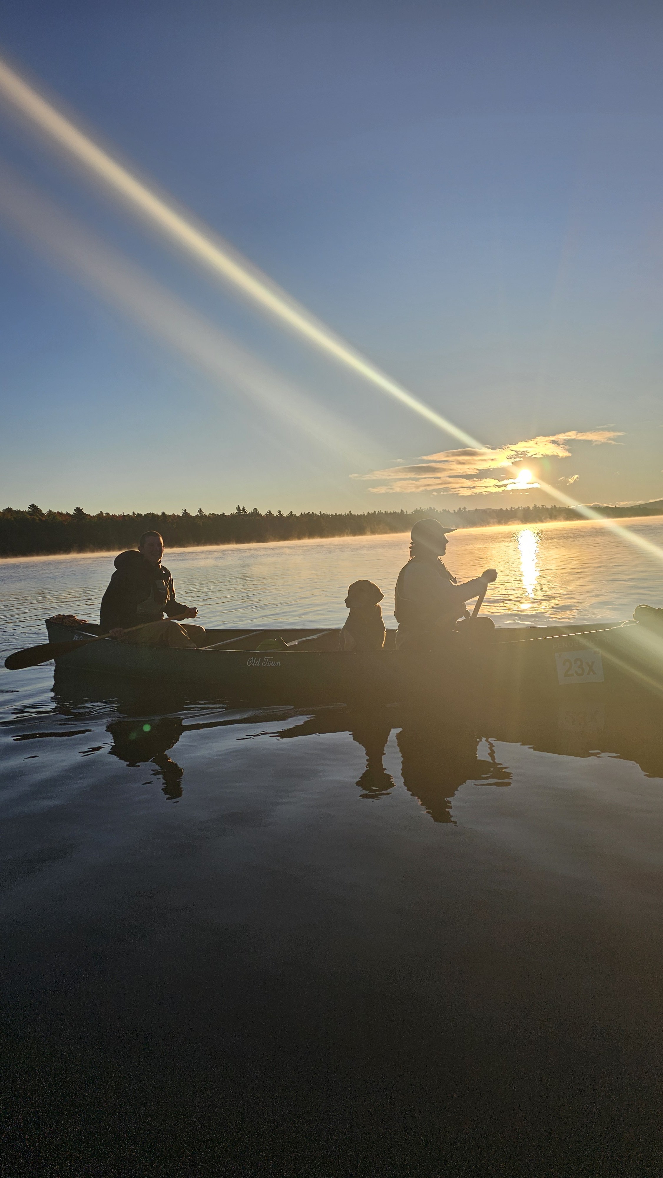 Flagstaff Lake, Maine. Two friends canoe with a dog on a calm lake at sunrise.